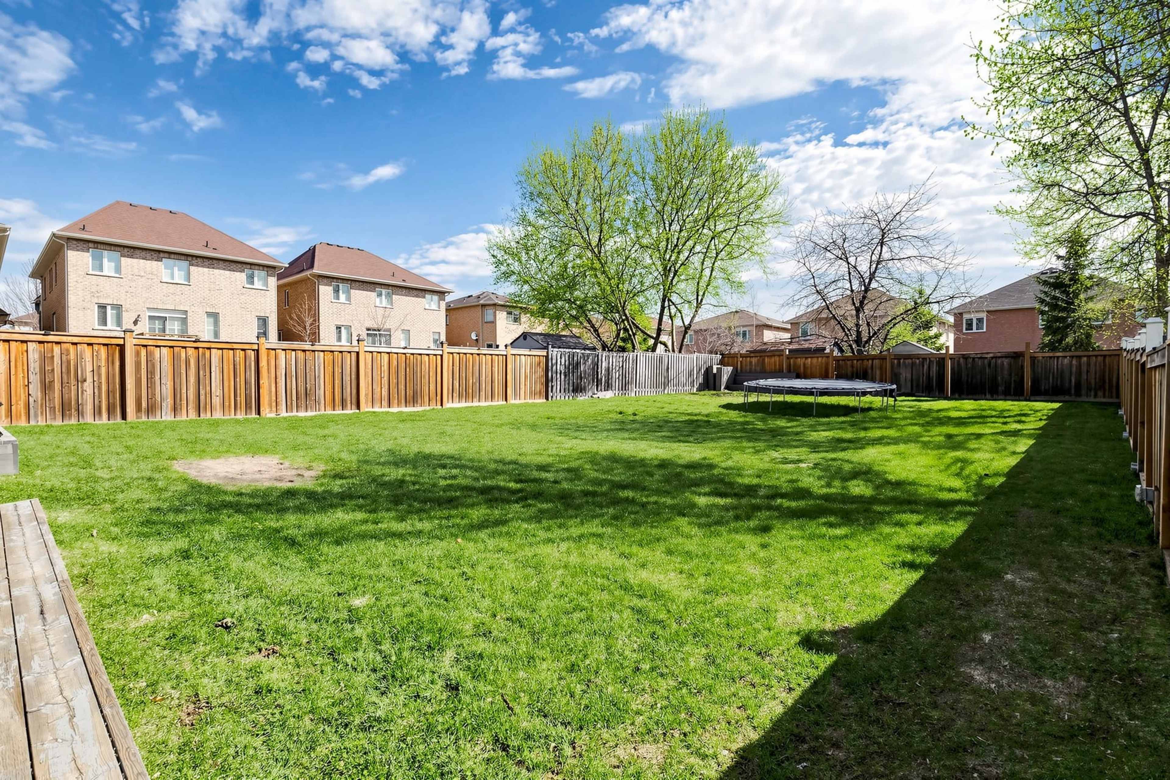The image shows a spacious grassy backyard enclosed by wooden fences, with two houses in the background and a trampoline partially visible on one side.