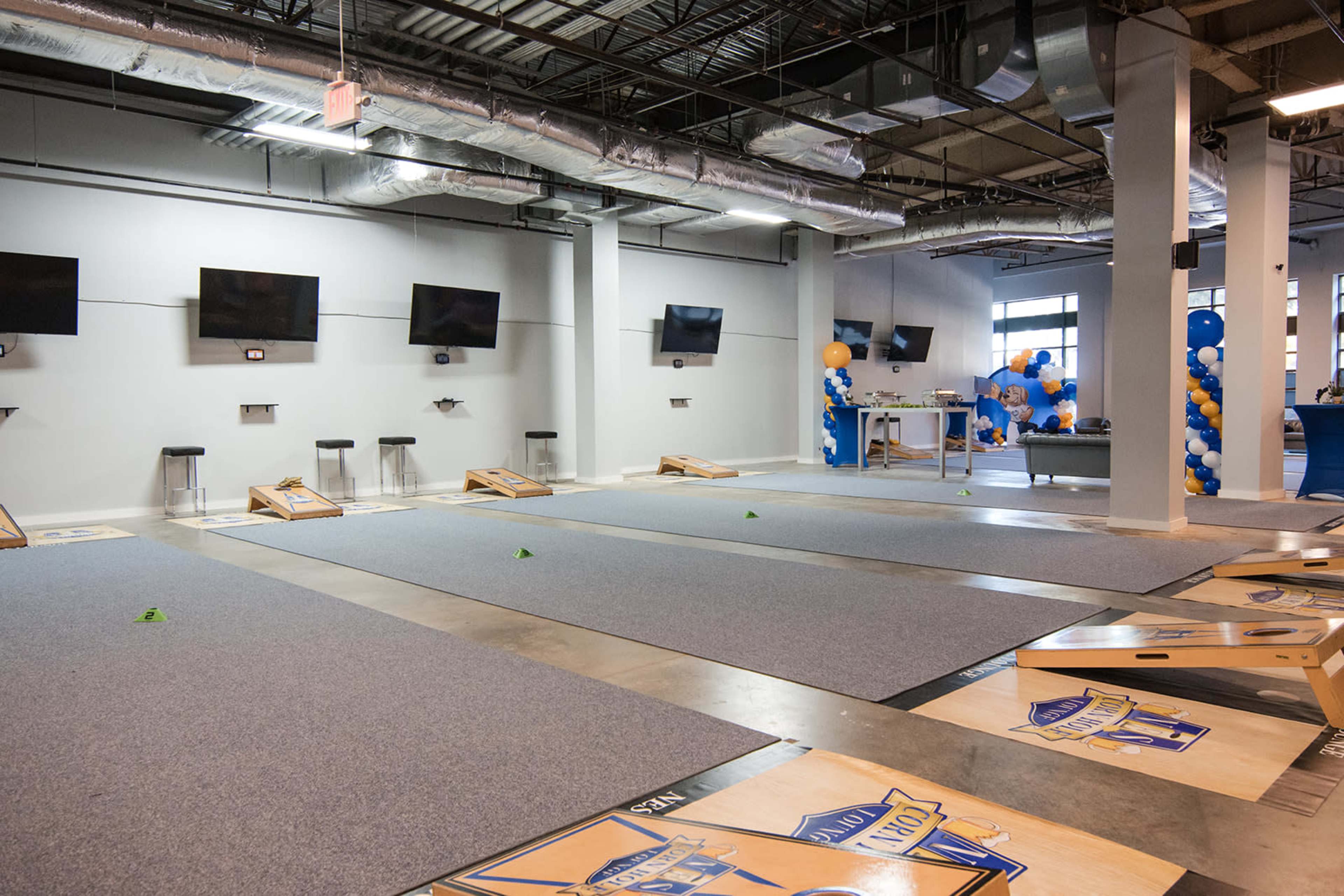 The image shows a spacious indoor area set up for cornhole games, featuring several cornhole boards on a carpeted floor and multiple screens mounted on the walls.