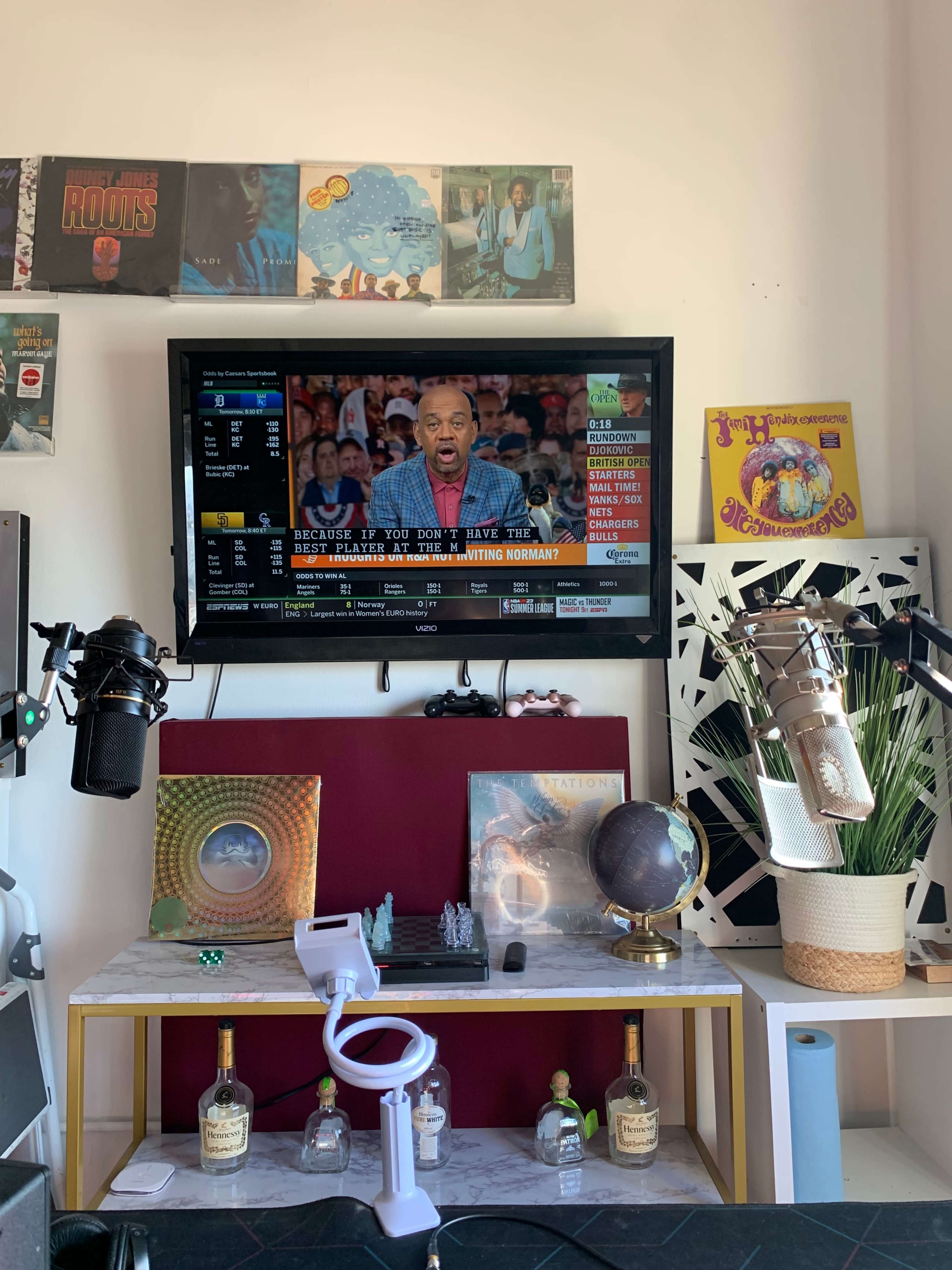 The scene features a television displaying a news program, placed above a table decorated with various objects, including microphones and bottles.