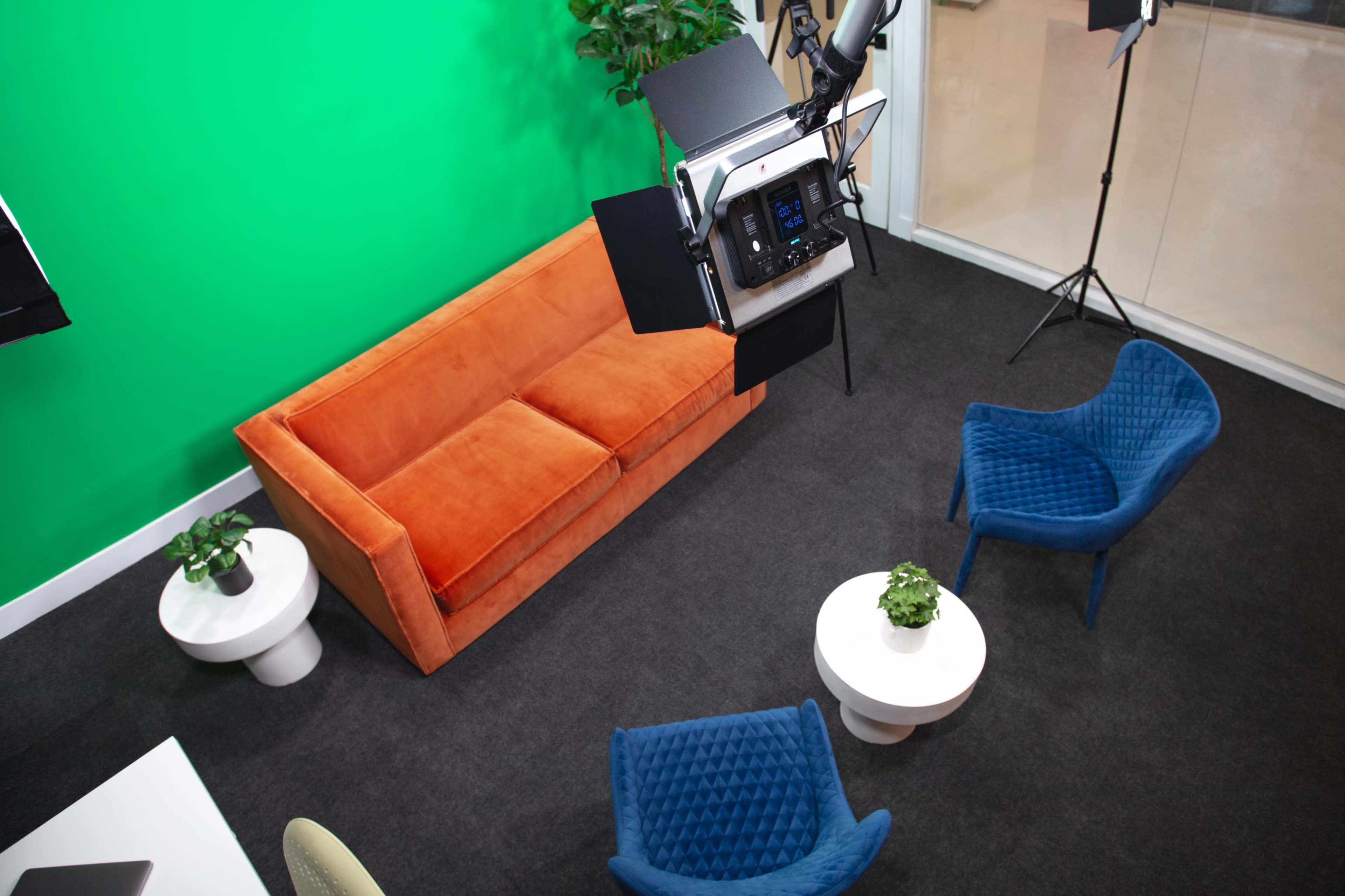 A modern studio setup with a green screen, an orange sofa, blue chairs, and a lighting rig overhead.