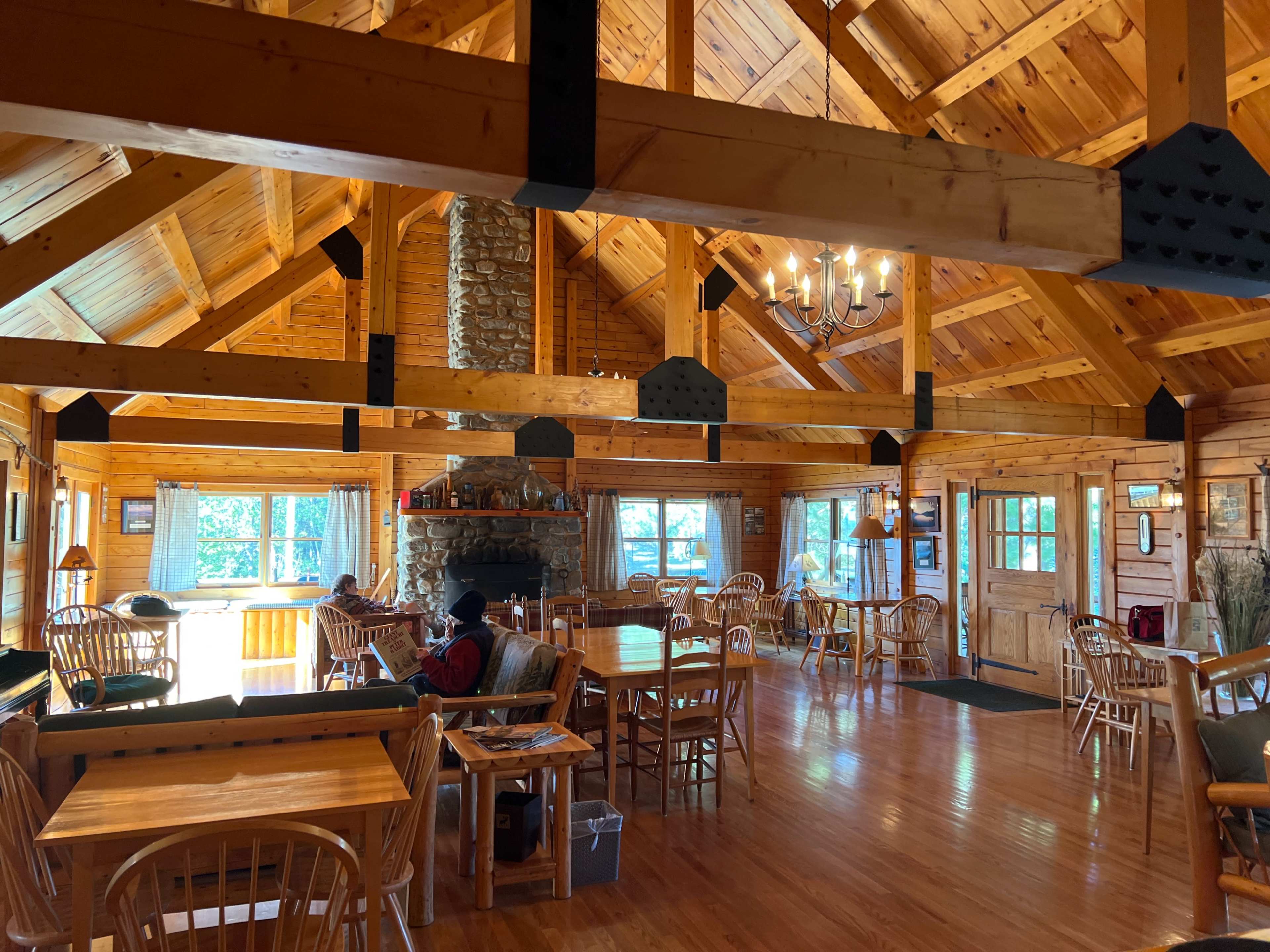 The interior of a rustic wooden lodge features a central stone fireplace, large windows, and wooden tables surrounded by chairs.
