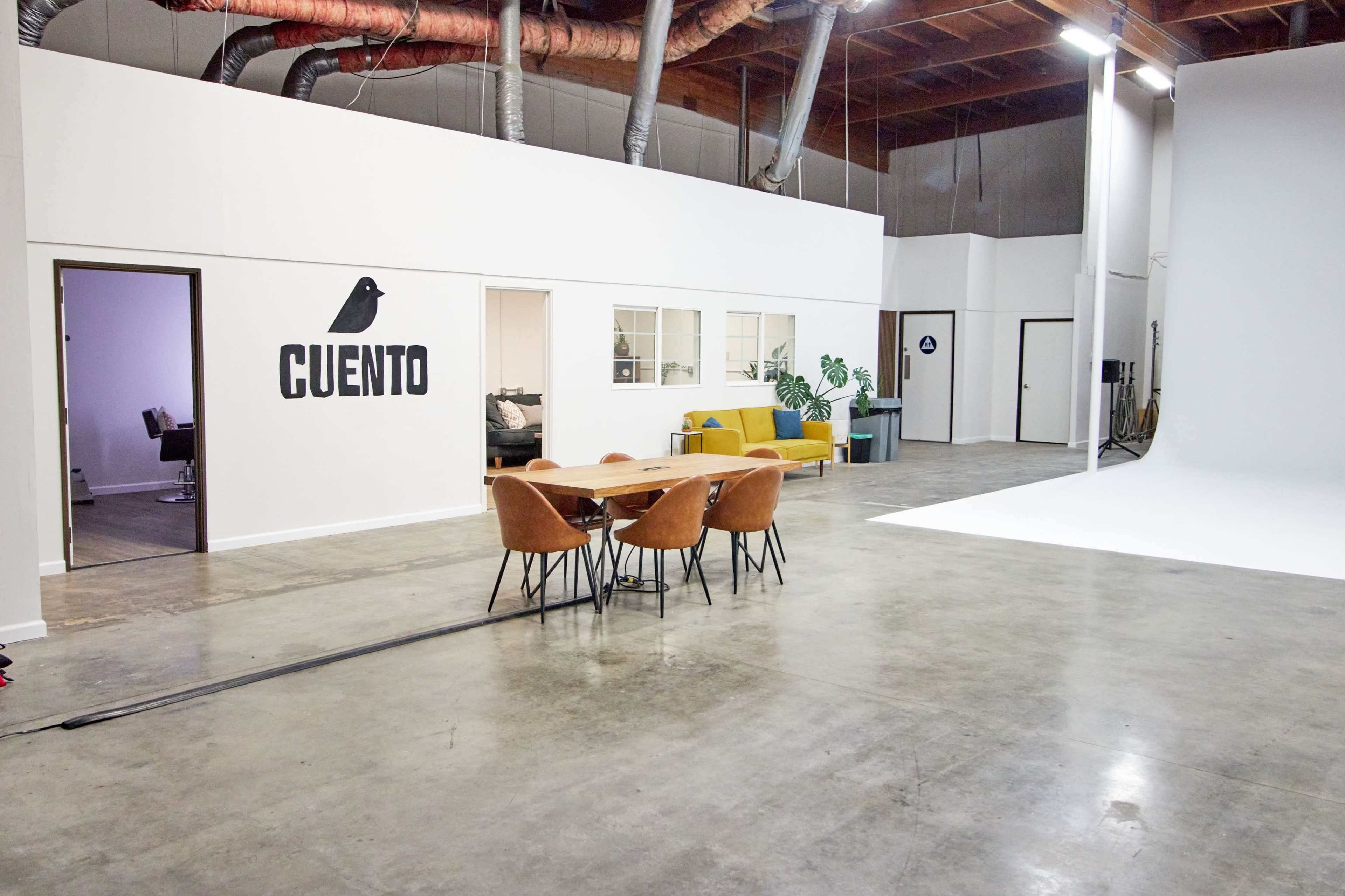 A modern studio space featuring a dining table with chairs, a sofa, and a white backdrop area, with the word "CUENTO" displayed on the wall.