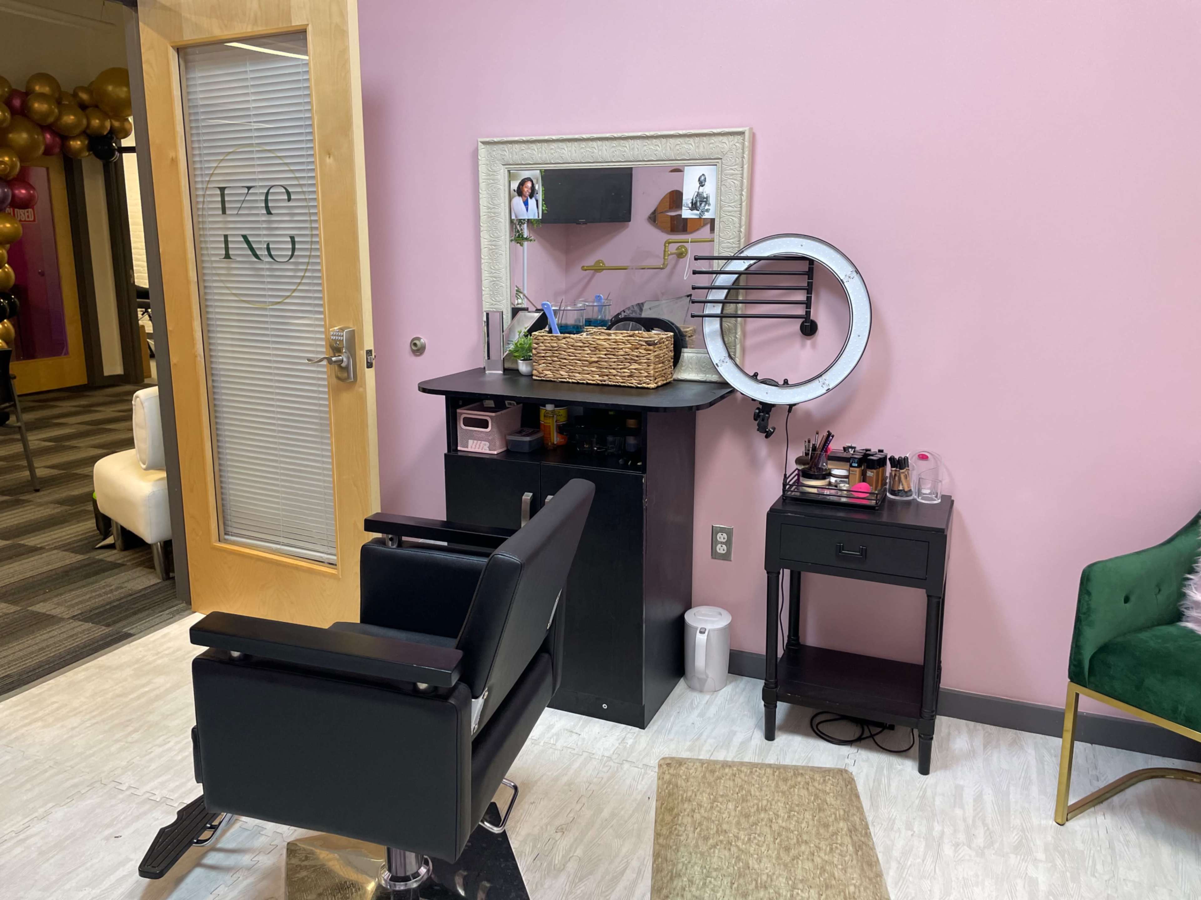 A salon corner features a black styling chair, a mirror above a dark cabinet, and a small table with various cosmetic items against a pink wall.
