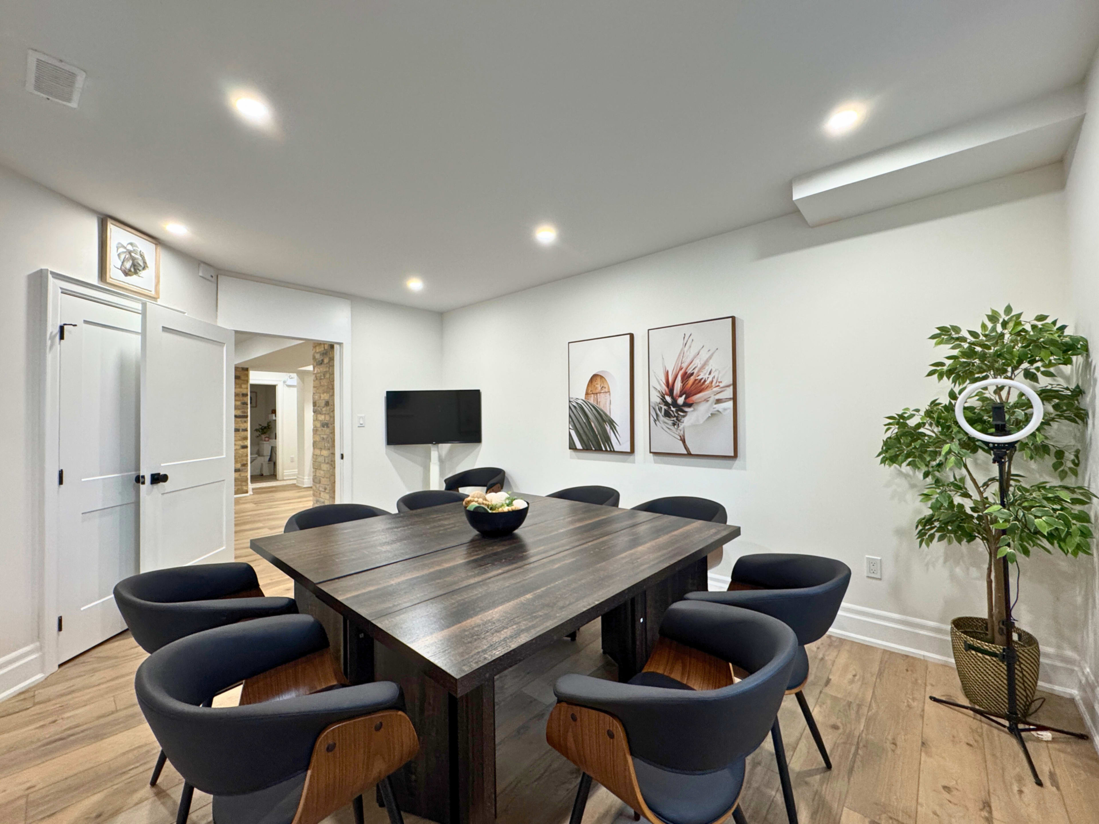 A modern meeting room features a large wooden table surrounded by black chairs, with artwork on the walls and a plant in the corner.