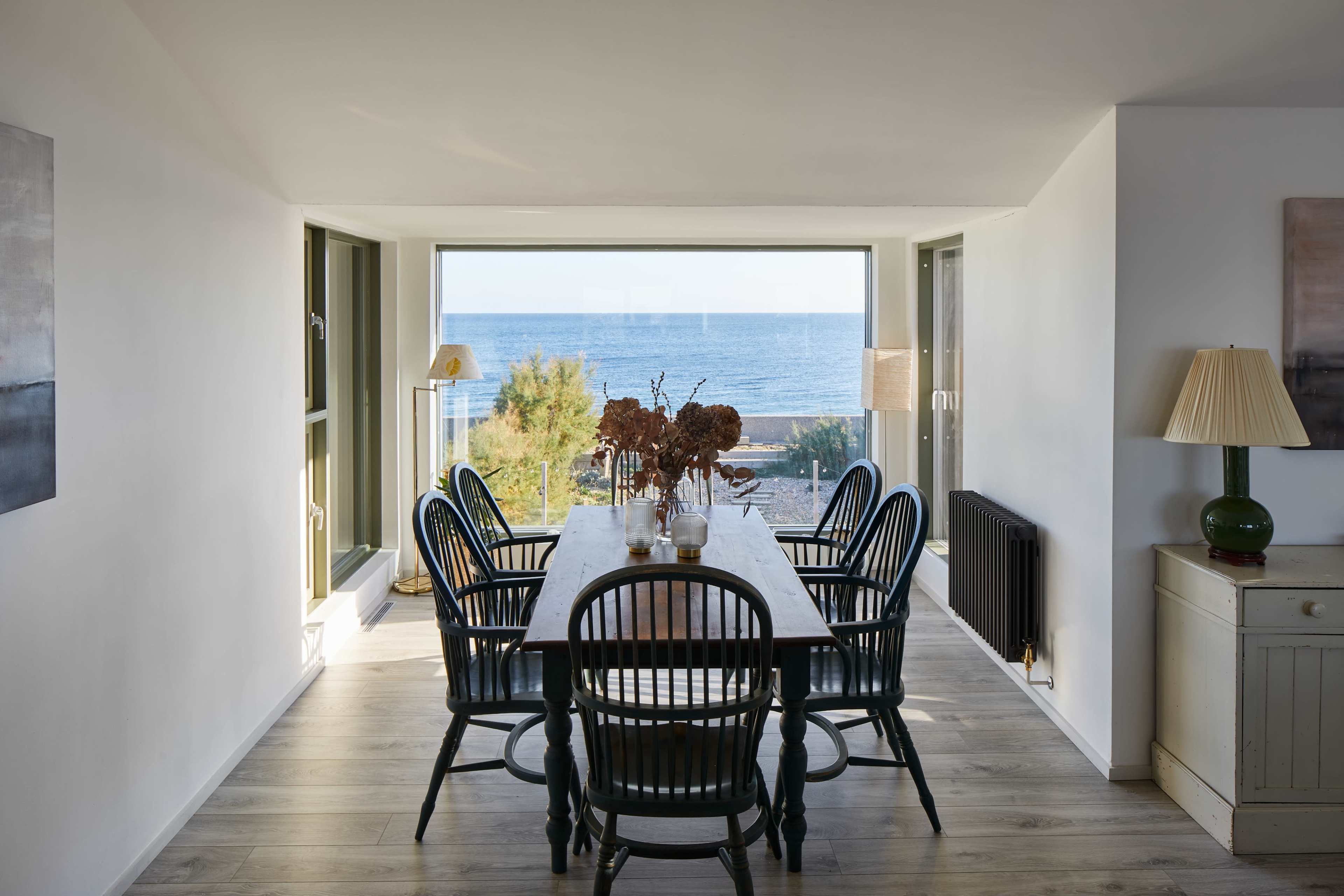 A dining room with a large table and six chairs overlooks a scenic view of the ocean through a wide window.