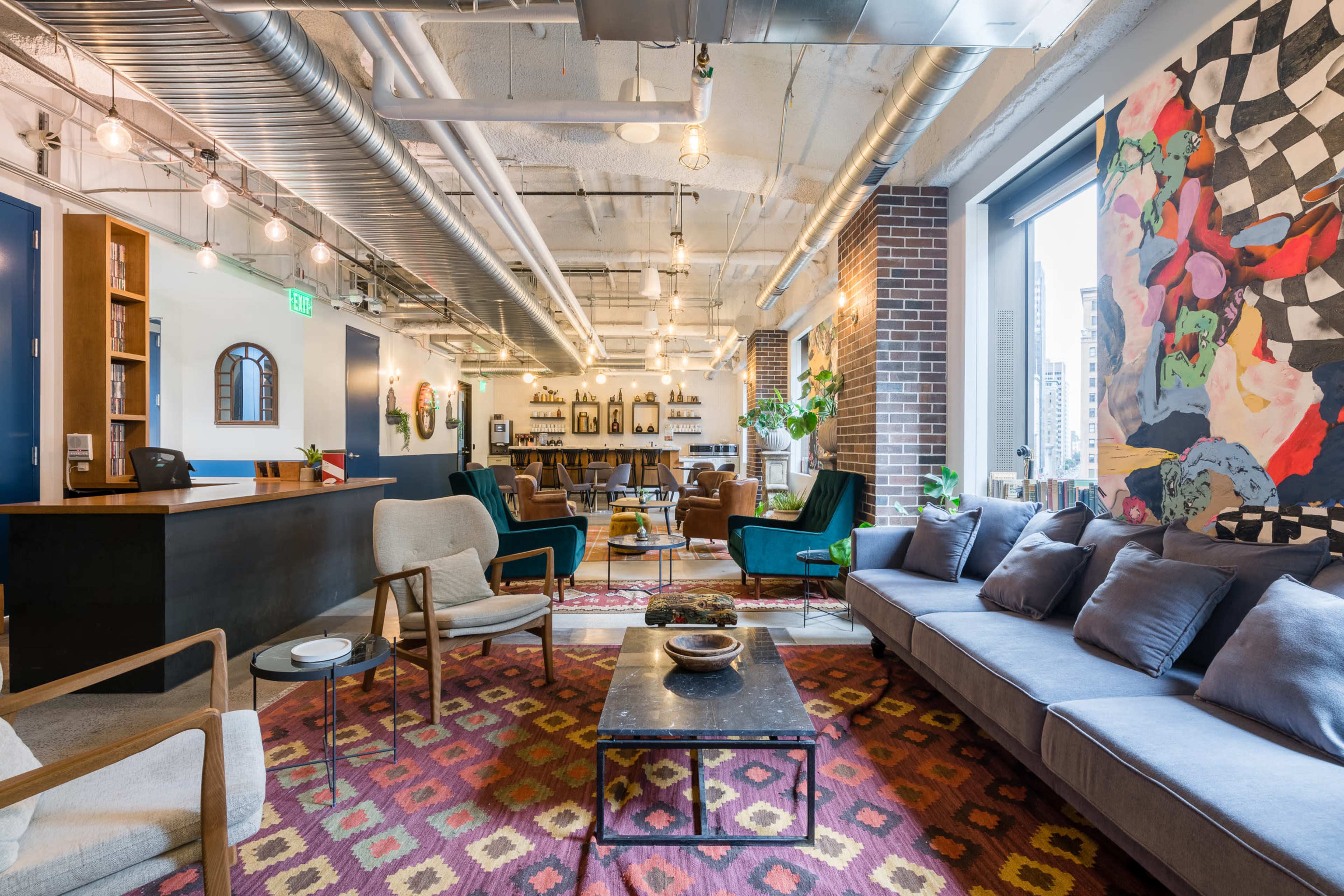 The image depicts a modern, brightly lit lounge area featuring comfortable seating arrangements, a decorative rug, and an artistic mural on the wall.