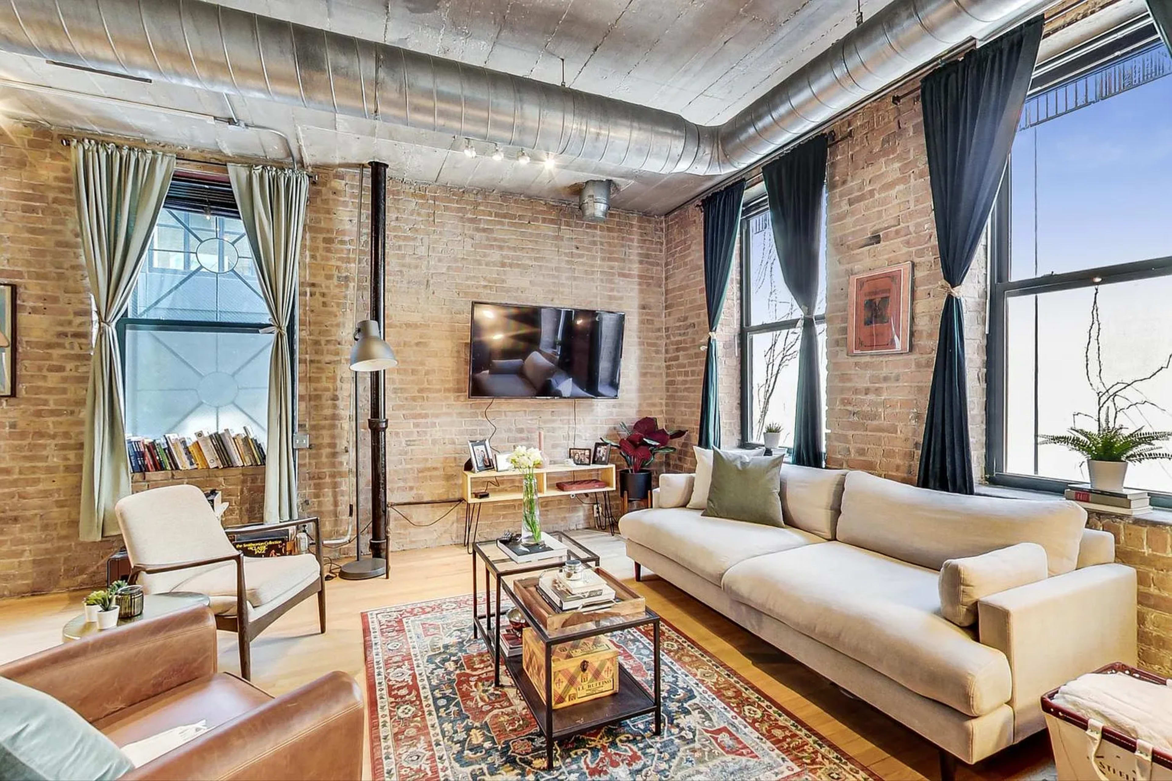 The image shows a stylish living room with exposed brick walls, a large television mounted on the wall, two windows, and a mix of modern and vintage furniture.
