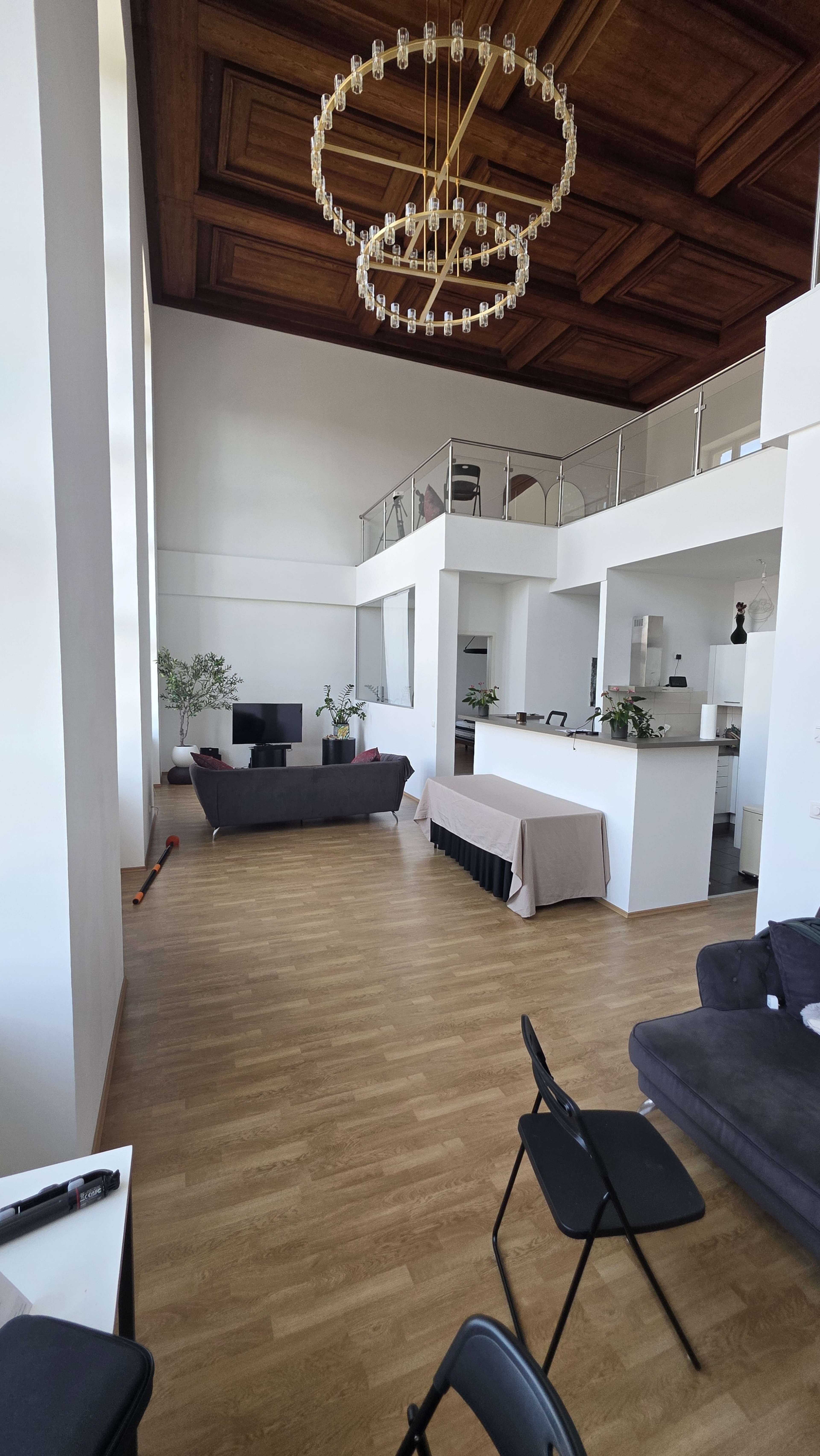 The image shows a spacious living area with high ceilings, a modern chandelier, and a partition separating a kitchen area from the lounge, furnished with a sofa and a couple of chairs.