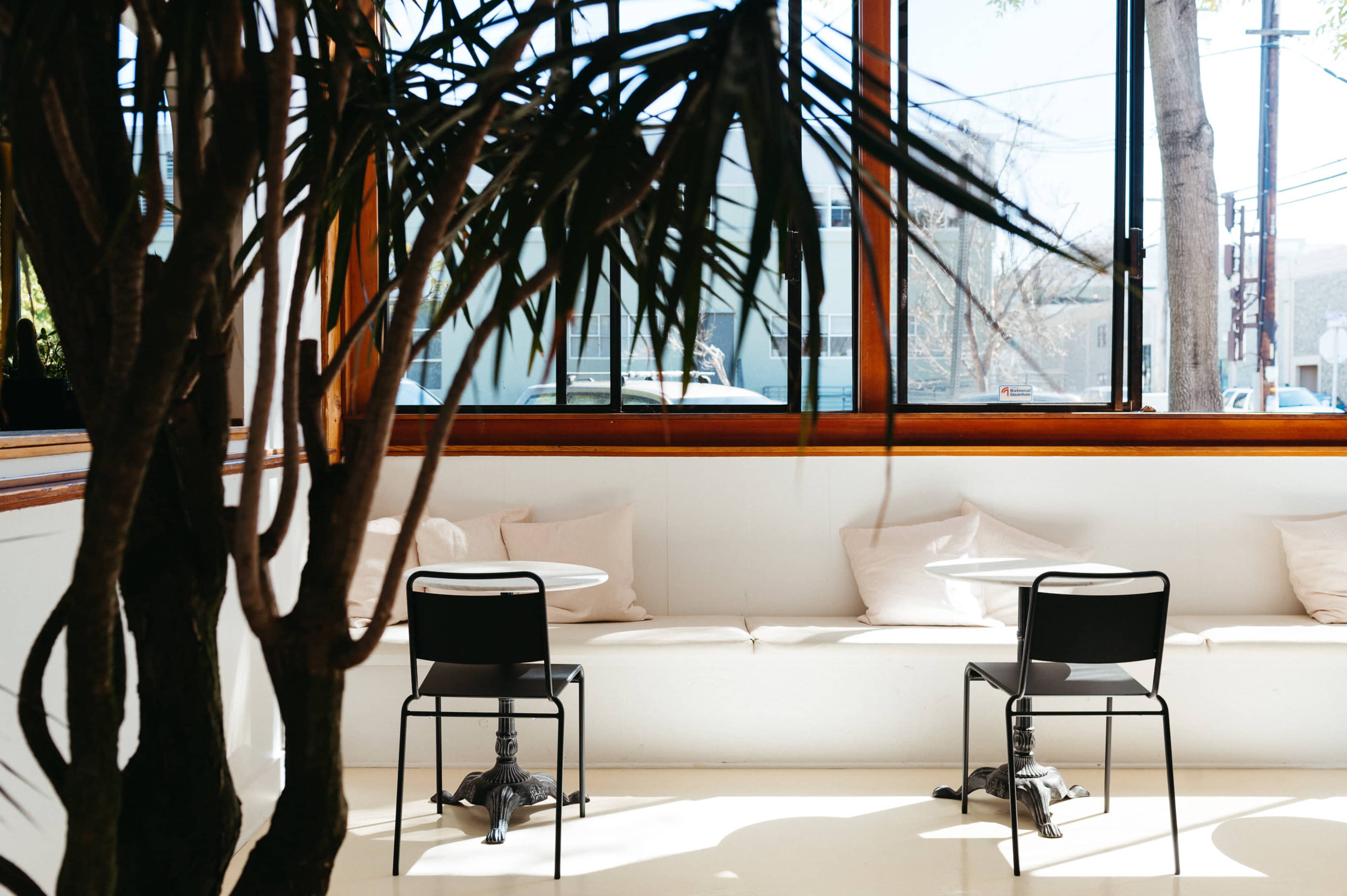 A sunlit café interior features two black chairs and small round tables near a window, with plush cushions on a white bench and a potted plant in the foreground.