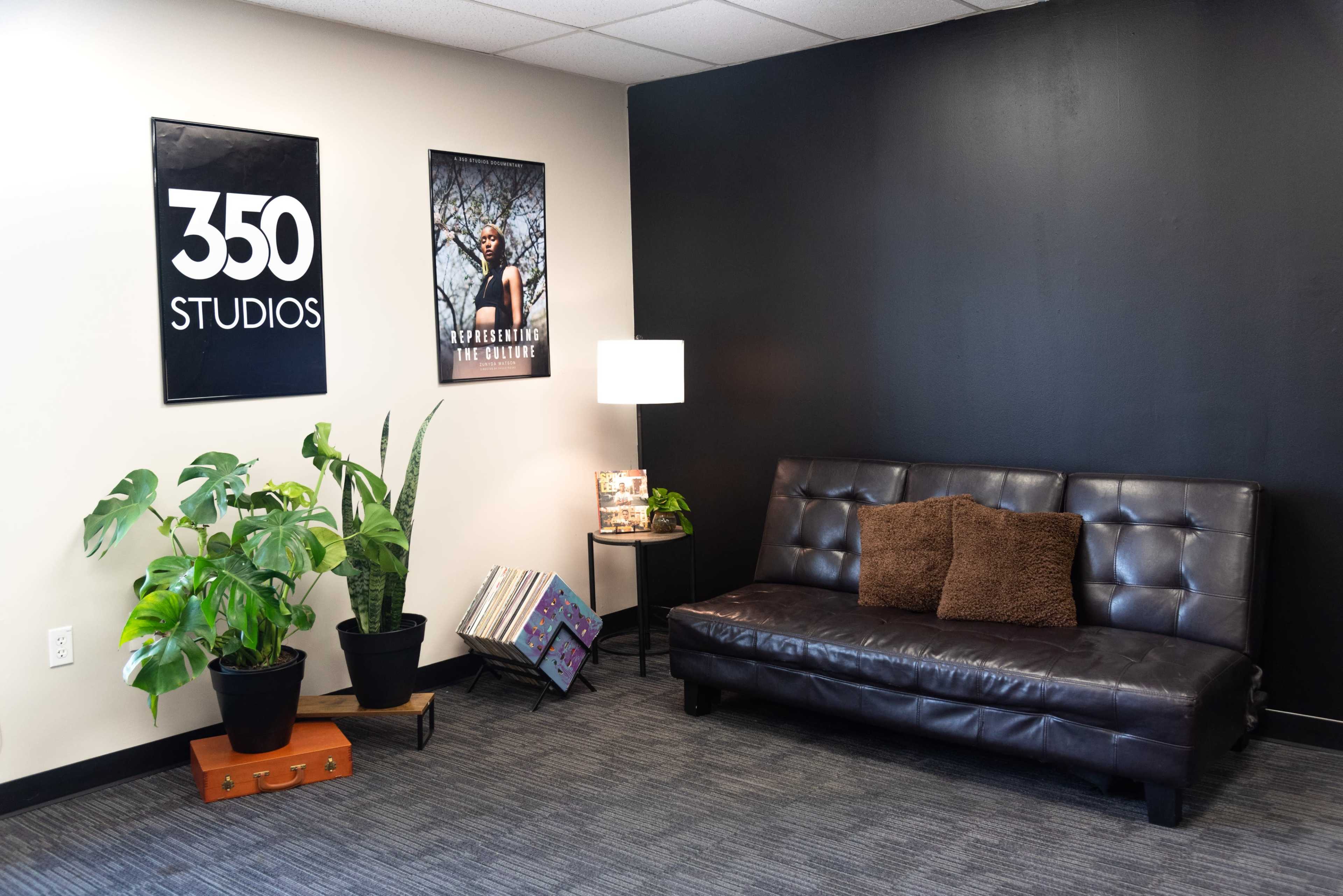A waiting area featuring a black wall, a brown sofa with two cushions, a lamp, plants, posters on the wall, and a small stack of records.