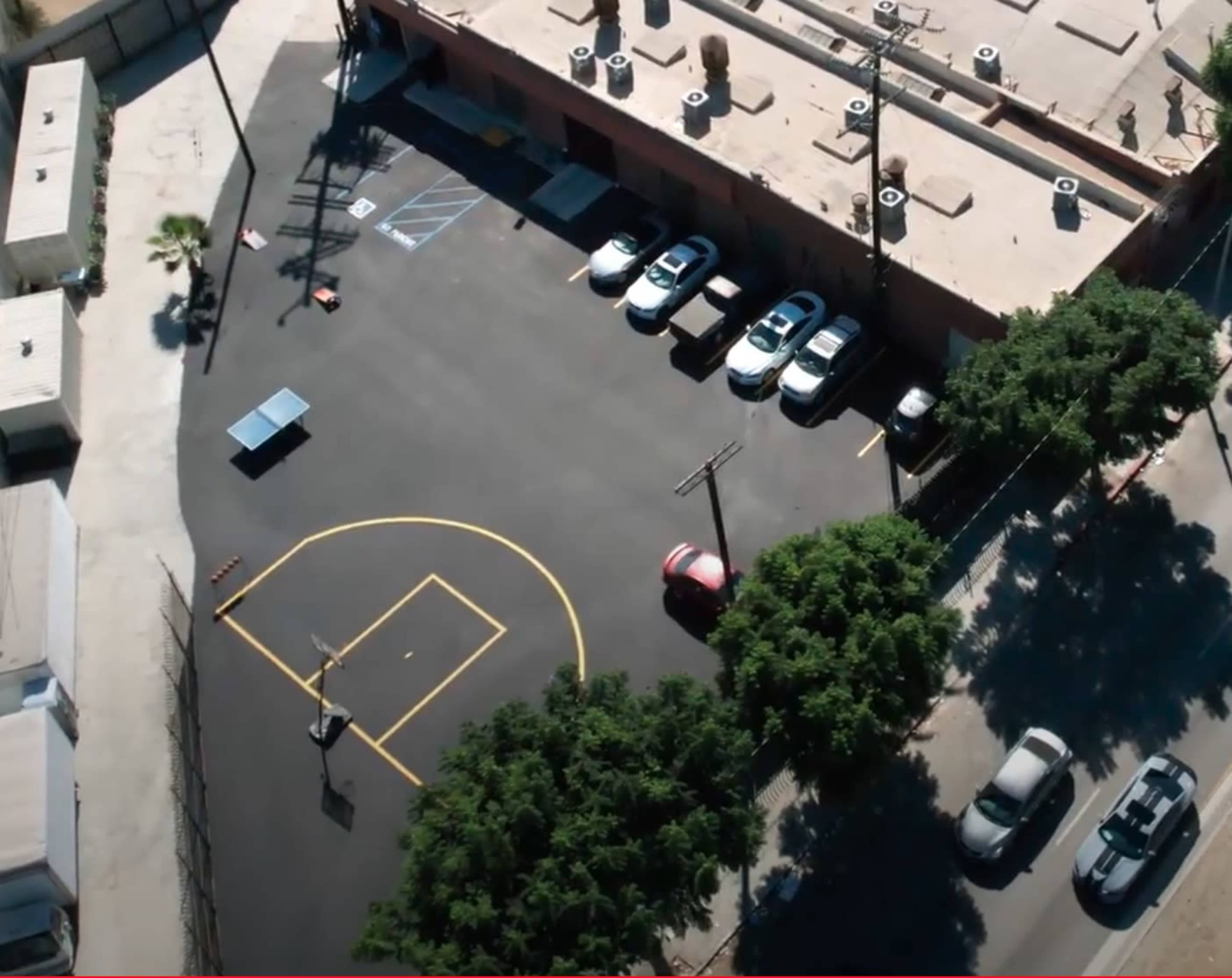 An aerial view of a parking lot with several cars, a basketball court marked in yellow, and surrounding trees.