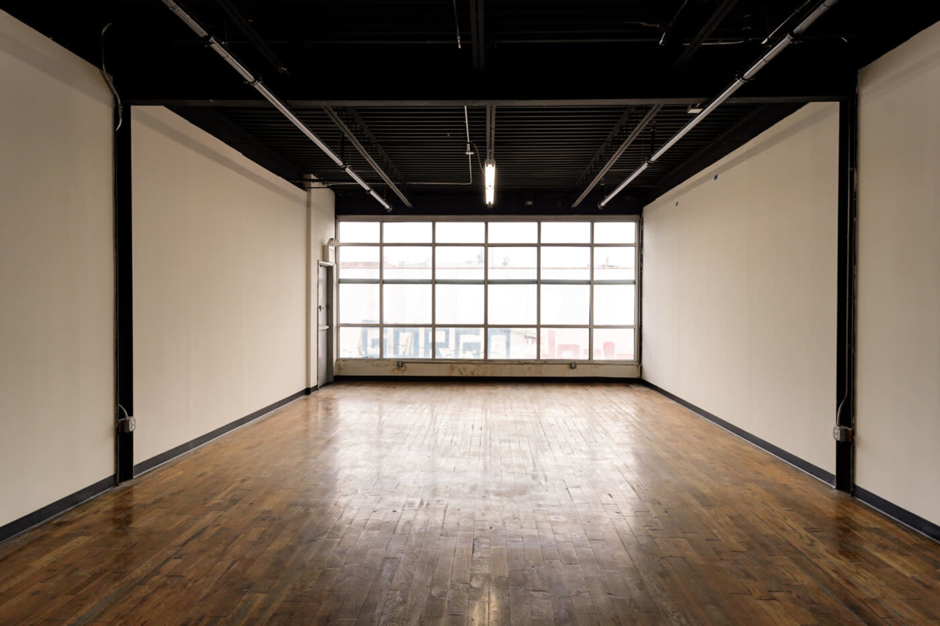 The image shows an empty room with wooden flooring and large windows along one wall, allowing natural light to enter the space.