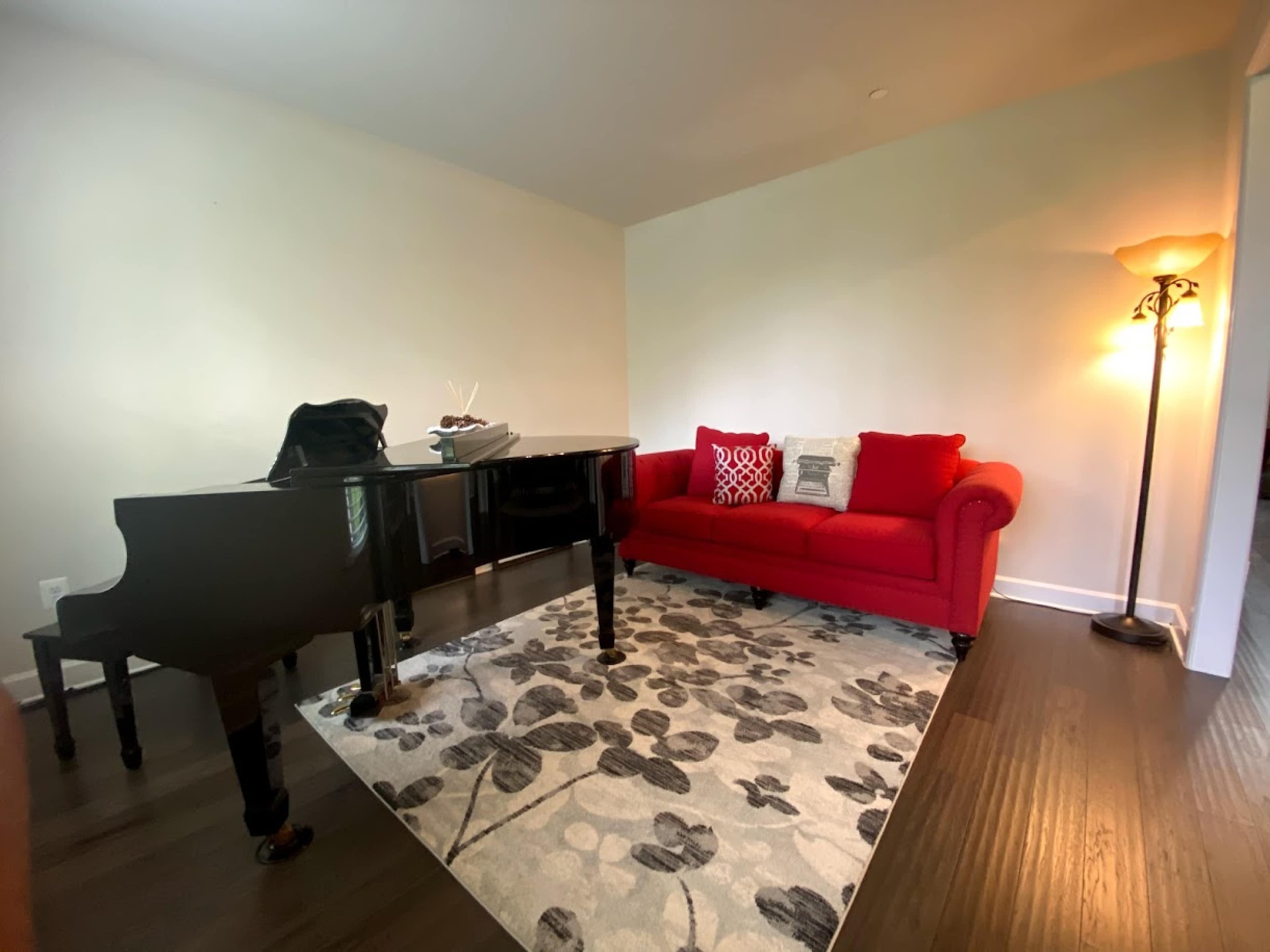 The image shows a room with a black grand piano, a red sofa with decorative pillows, and a standing lamp on a floral rug.
