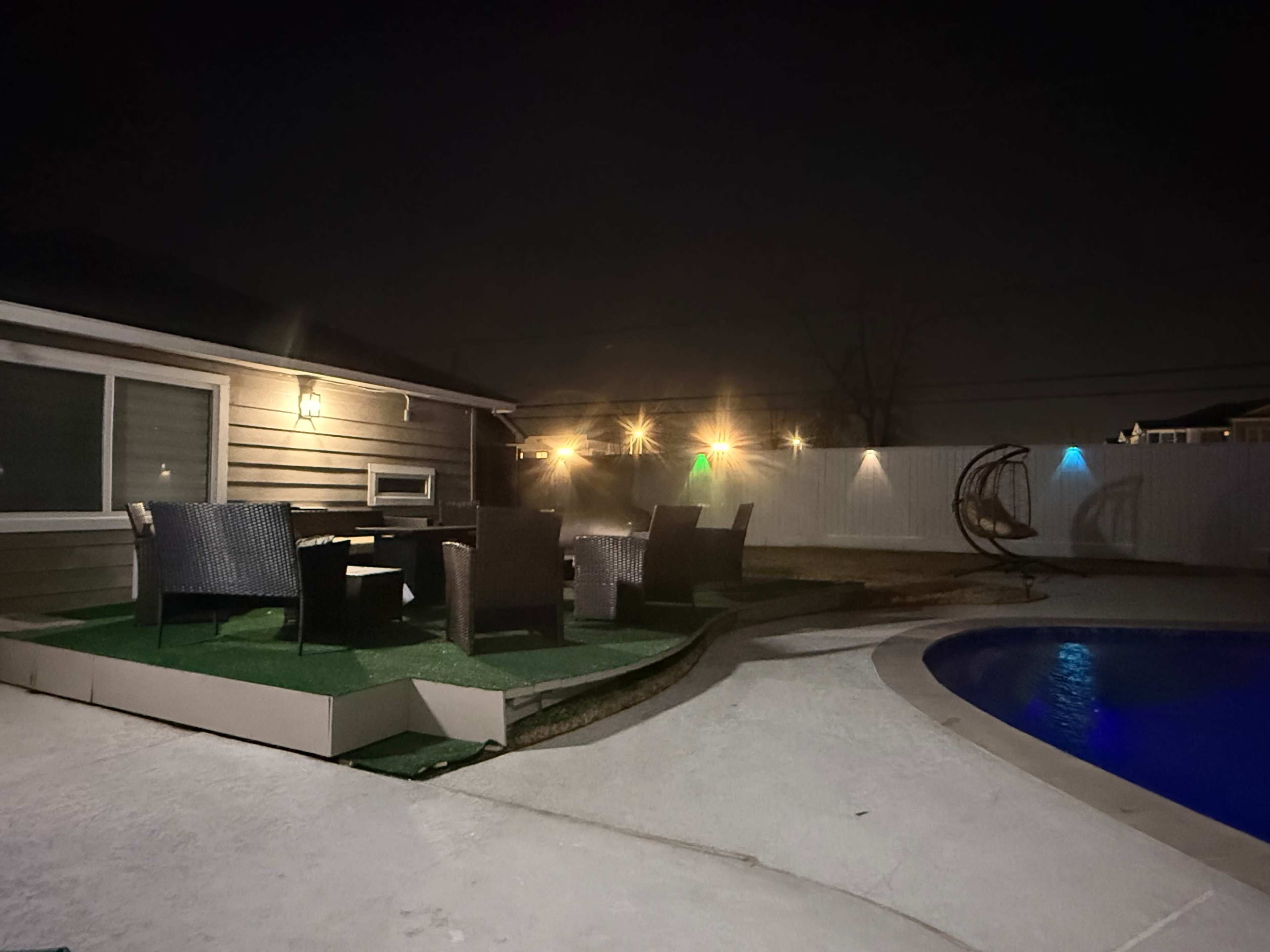 The image shows a backyard at night featuring a pool, a small green area with seating, and string lights illuminating the space.