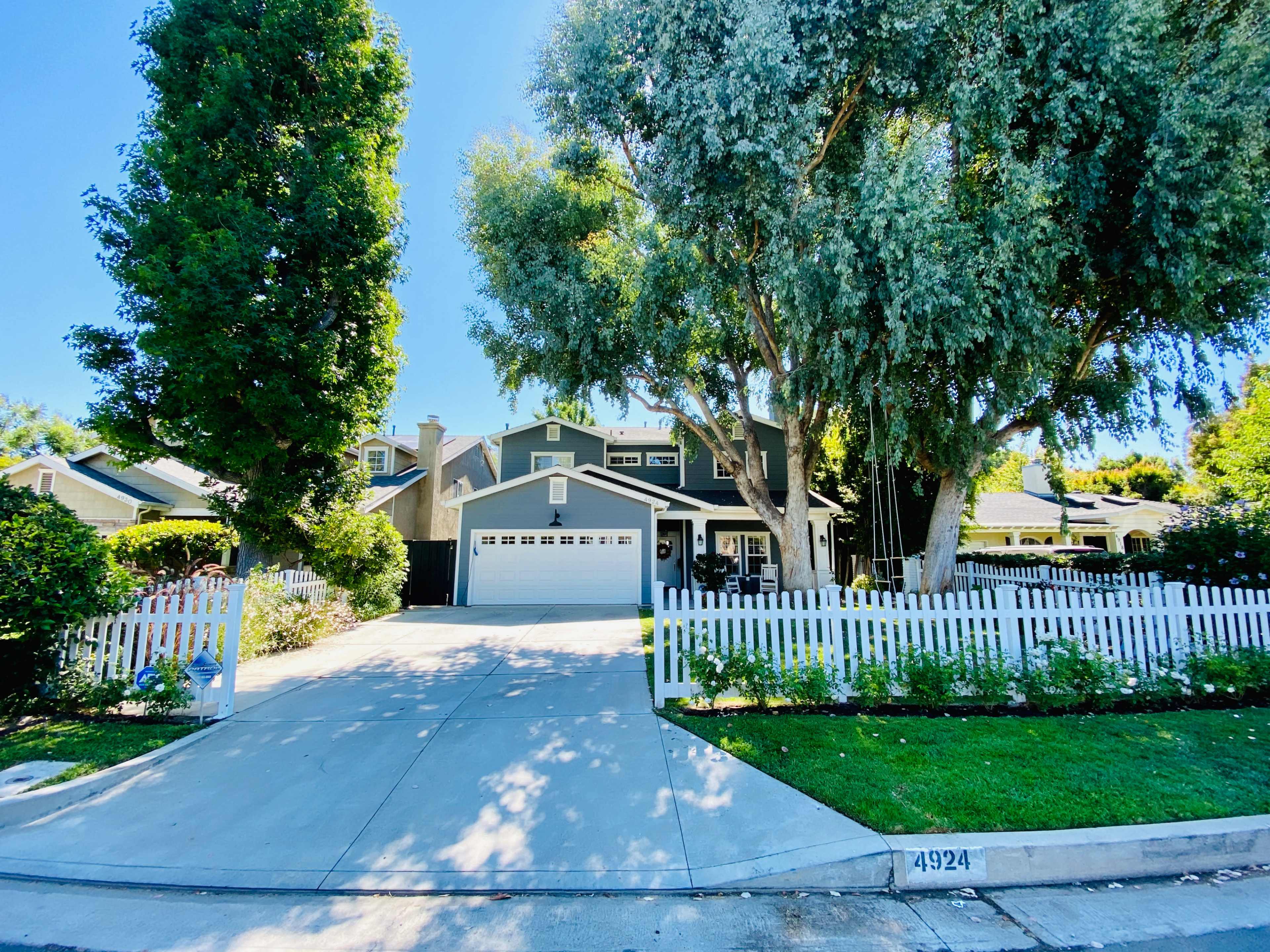 A two-story house with a front yard enclosed by a white picket fence, surrounded by large trees and a paved driveway.