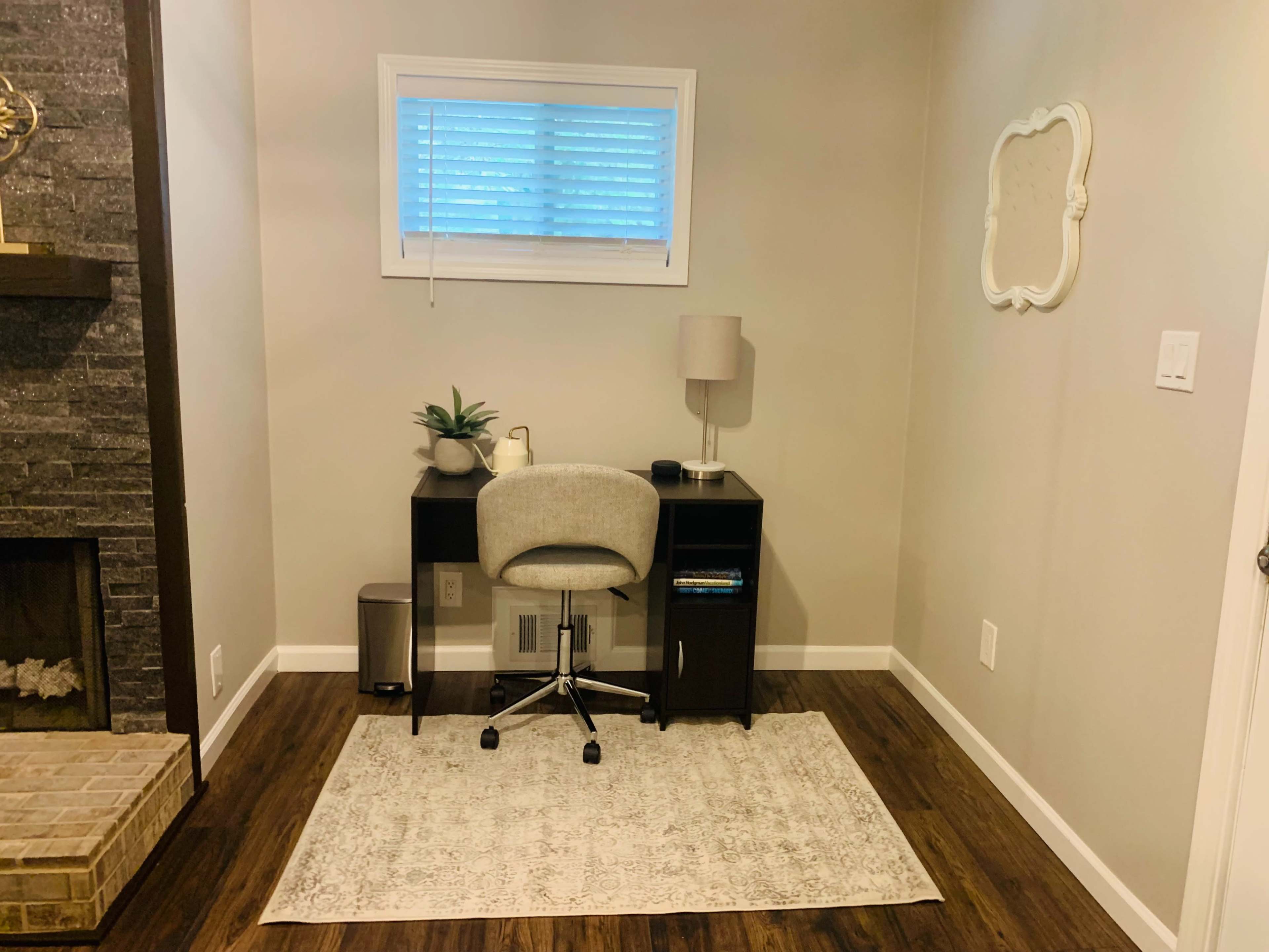 A small home office features a desk with a chair, a lamp, a plant, and a wastebasket, set against a light-colored wall and a window.