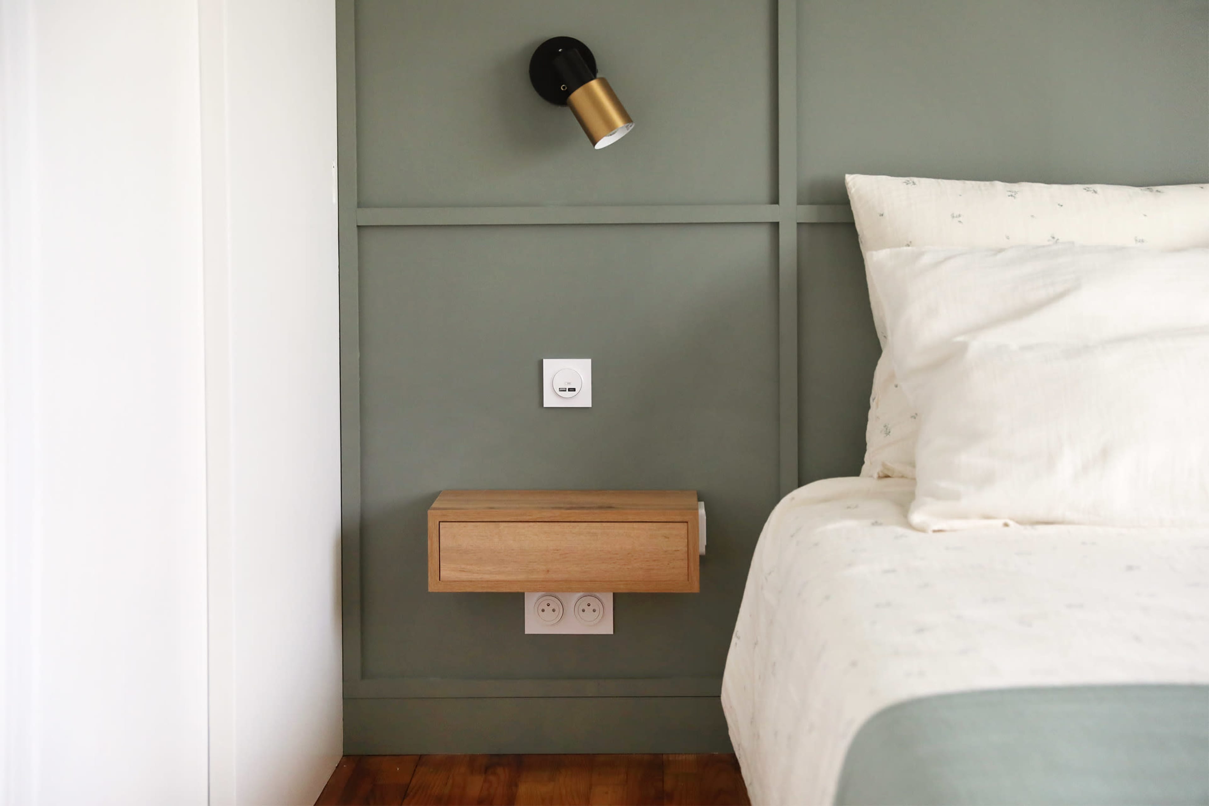 The image shows a minimalist bedroom corner featuring a light fixture above a wooden bedside table, which is mounted on a green wall with electrical outlets below it.