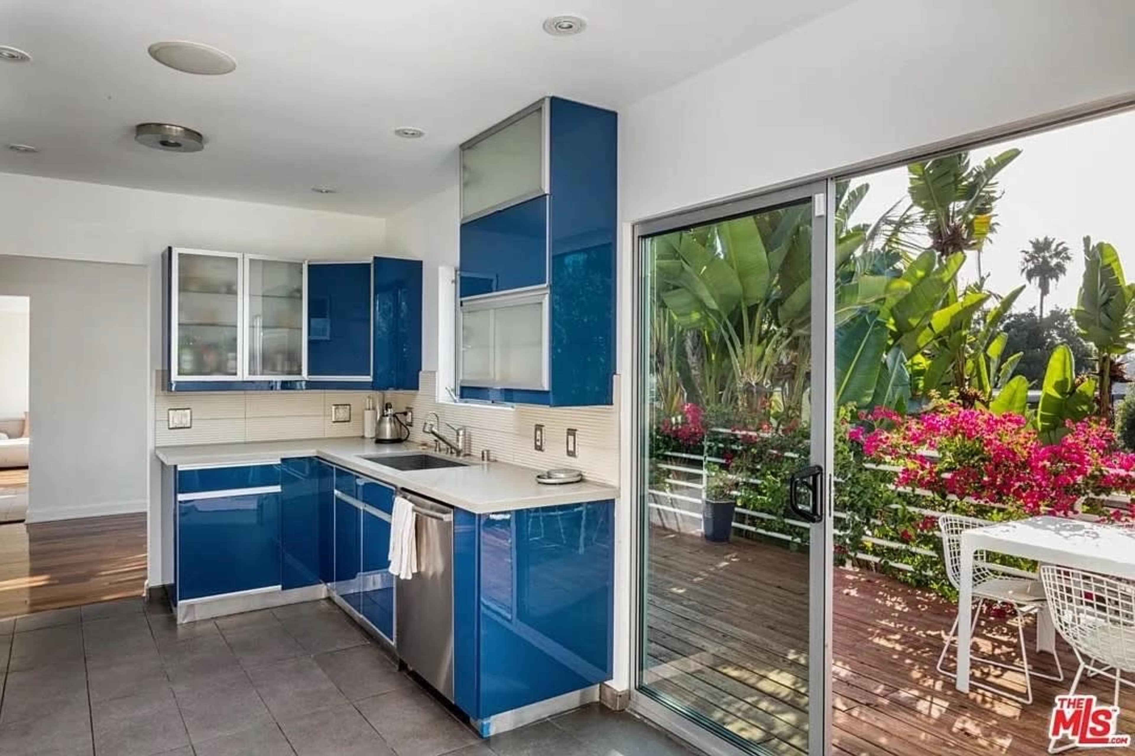 The image shows a modern kitchen with blue cabinets, a white countertop, and large windows overlooking a deck with greenery and flowers.