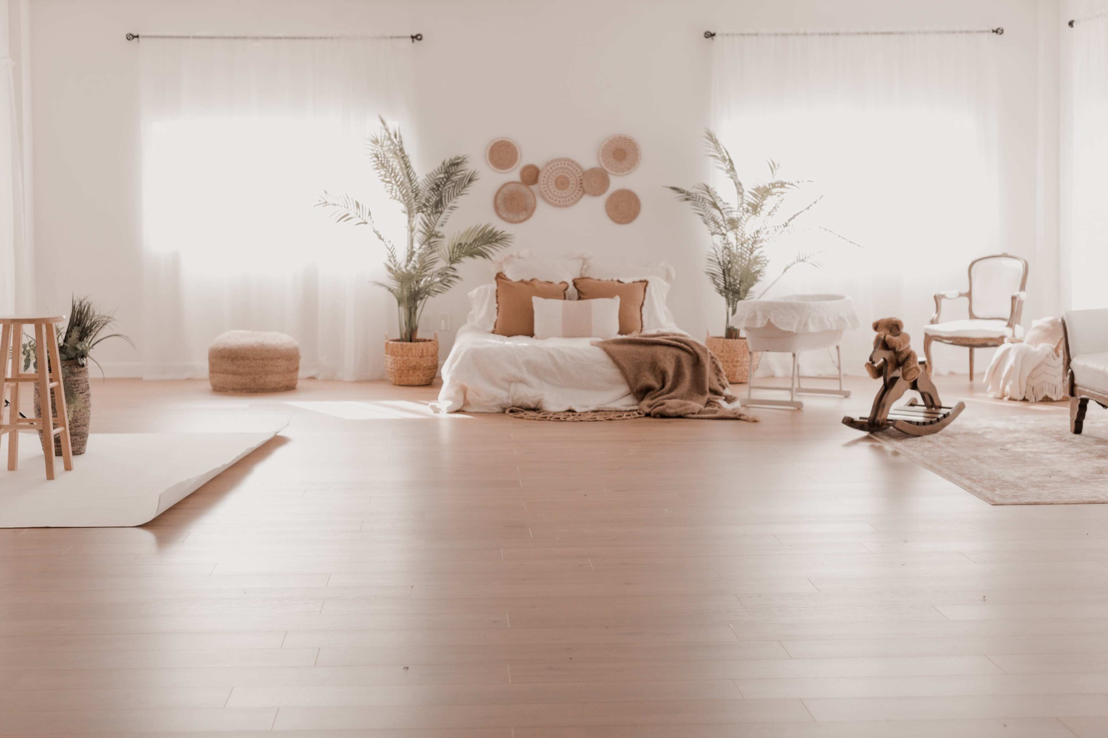 A bright and airy room features a bed with decorative pillows, plants in woven baskets, a rocking horse, and large windows letting in natural light.