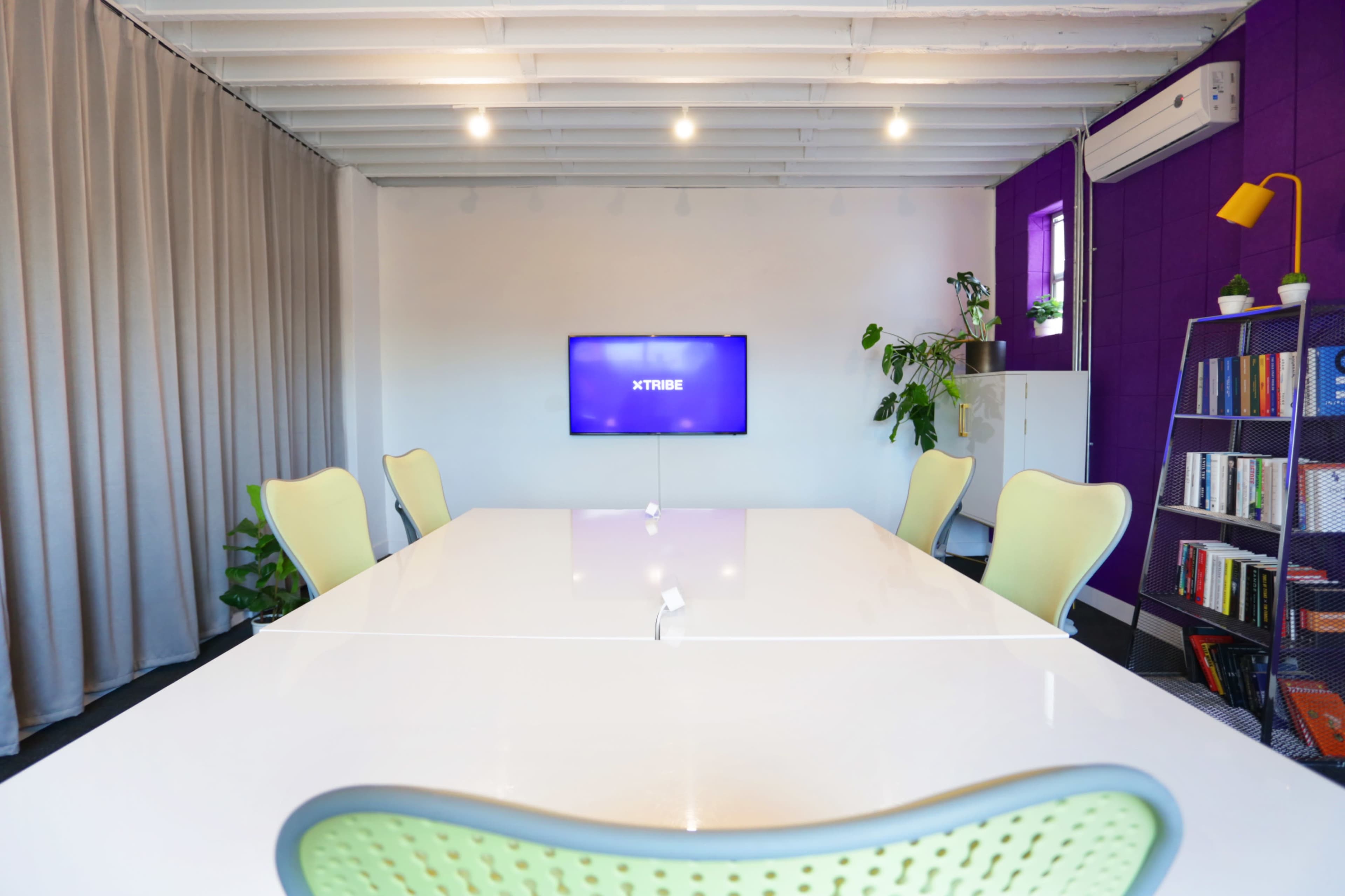 A conference room features a white table, green chairs, a television mounted on a purple wall, and plants along the sides.