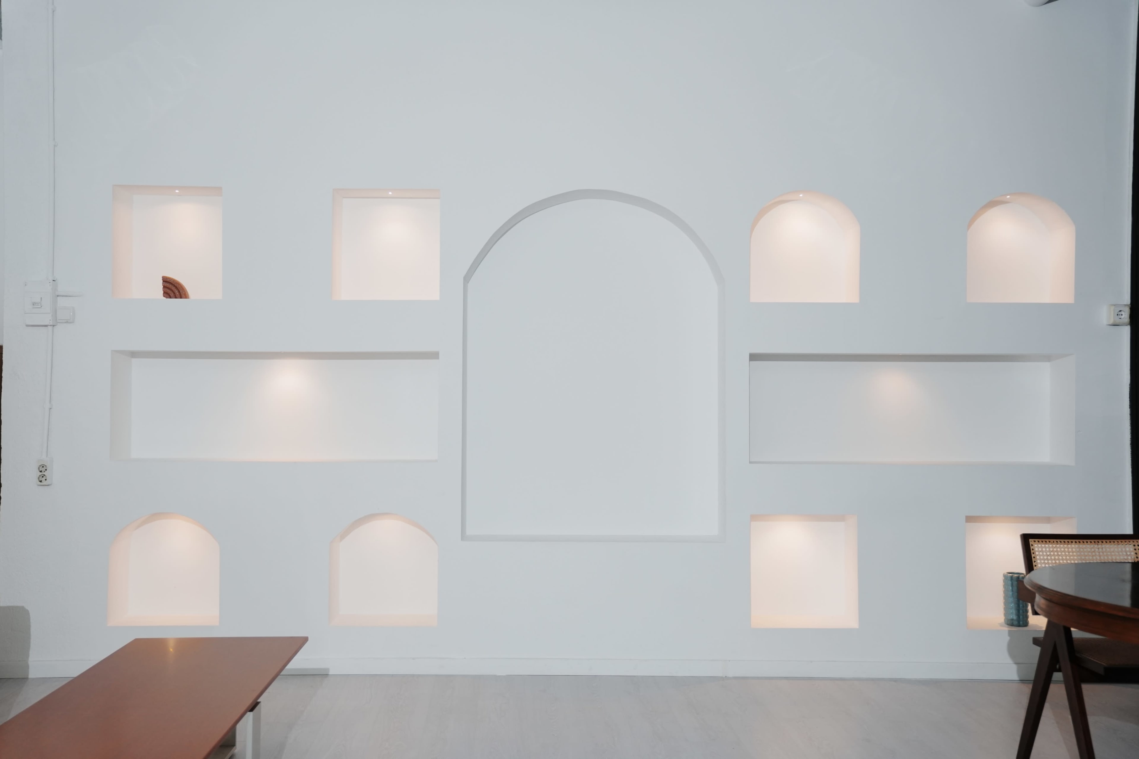 The image shows a white wall with several recessed alcoves, some illuminated, and a simple wooden table in the foreground.