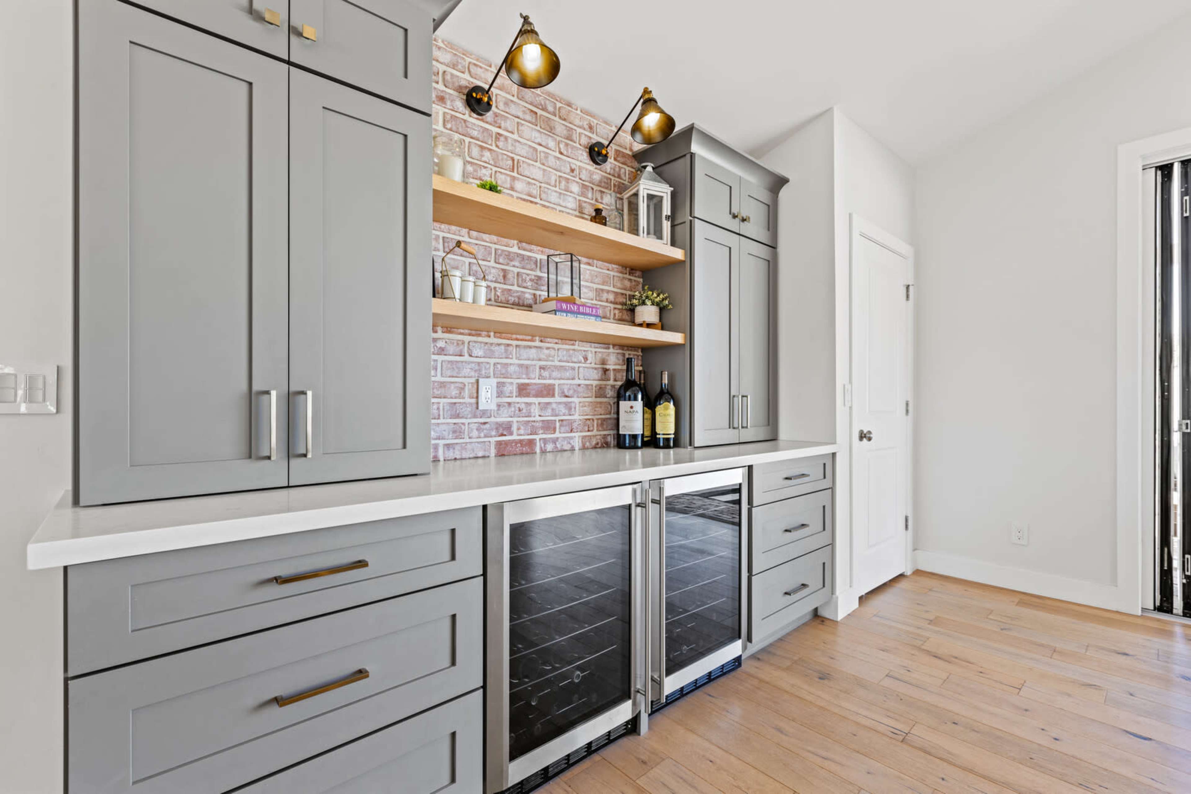 The image shows a modern kitchen bar area with gray cabinets, a white countertop, and open wooden shelves above a brick accent wall.