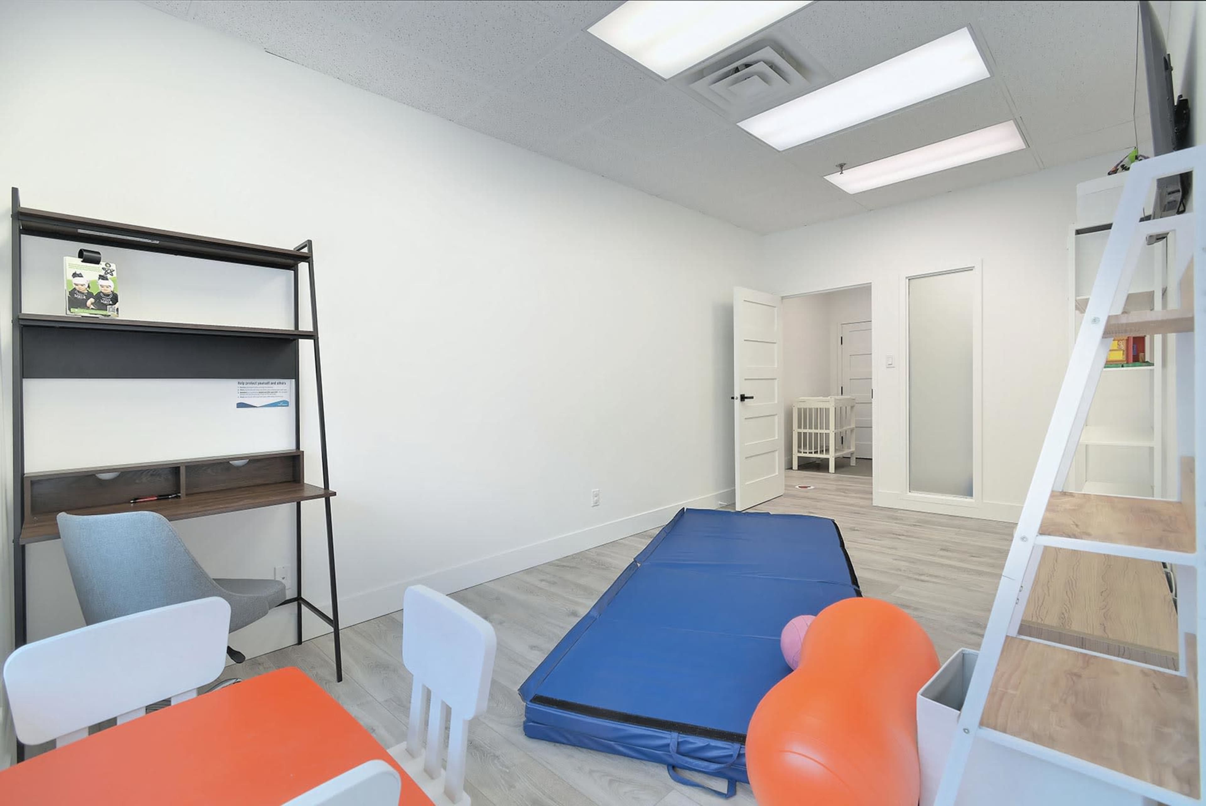A bright, minimalist room with a blue mat on the floor, a small orange exercise ball, a wooden bookshelf, a chair, and a white table and chairs.