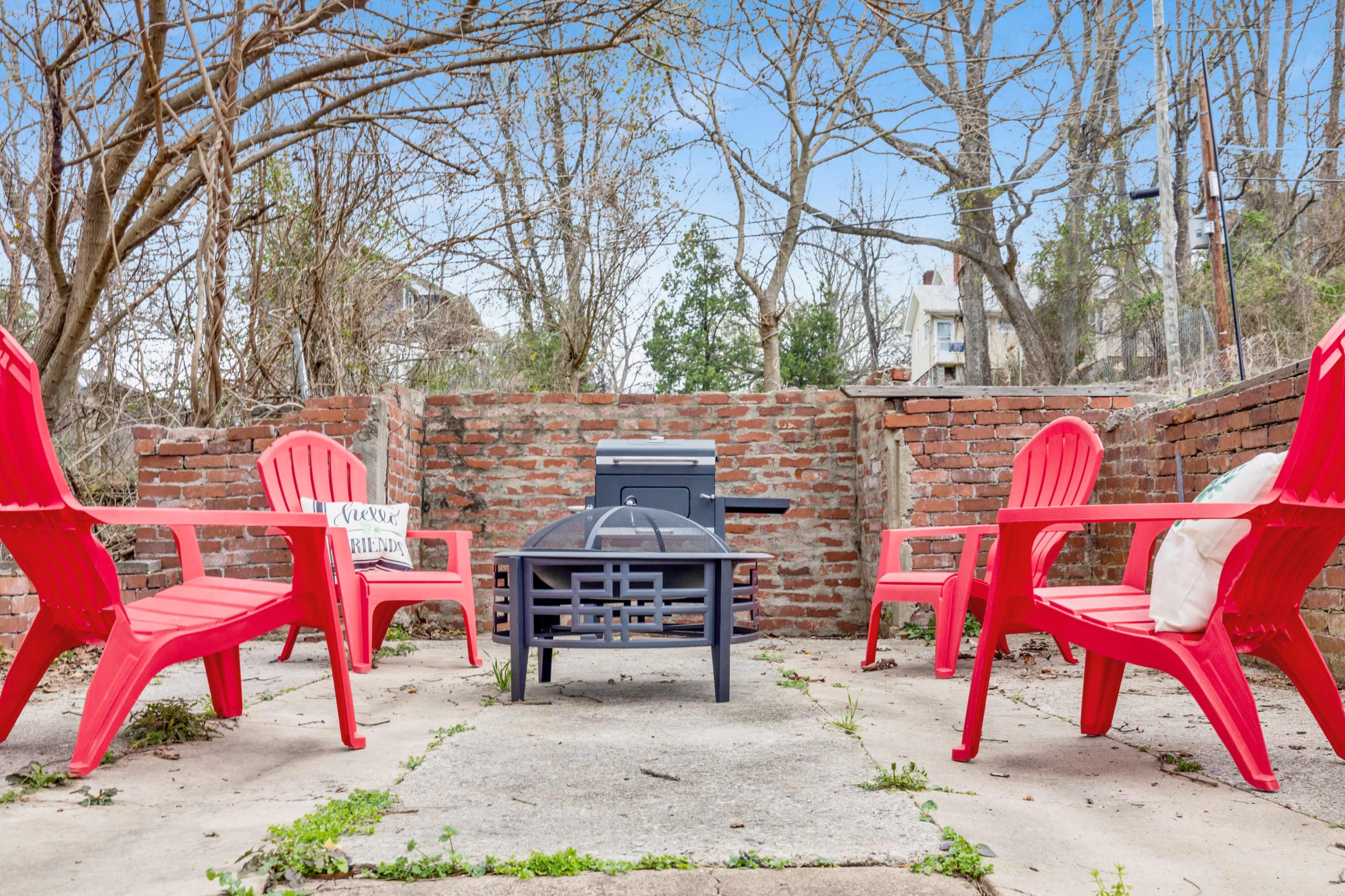 A small outdoor seating area features four red chairs arranged around a central concrete surface, with a grill and brick walls in the background.