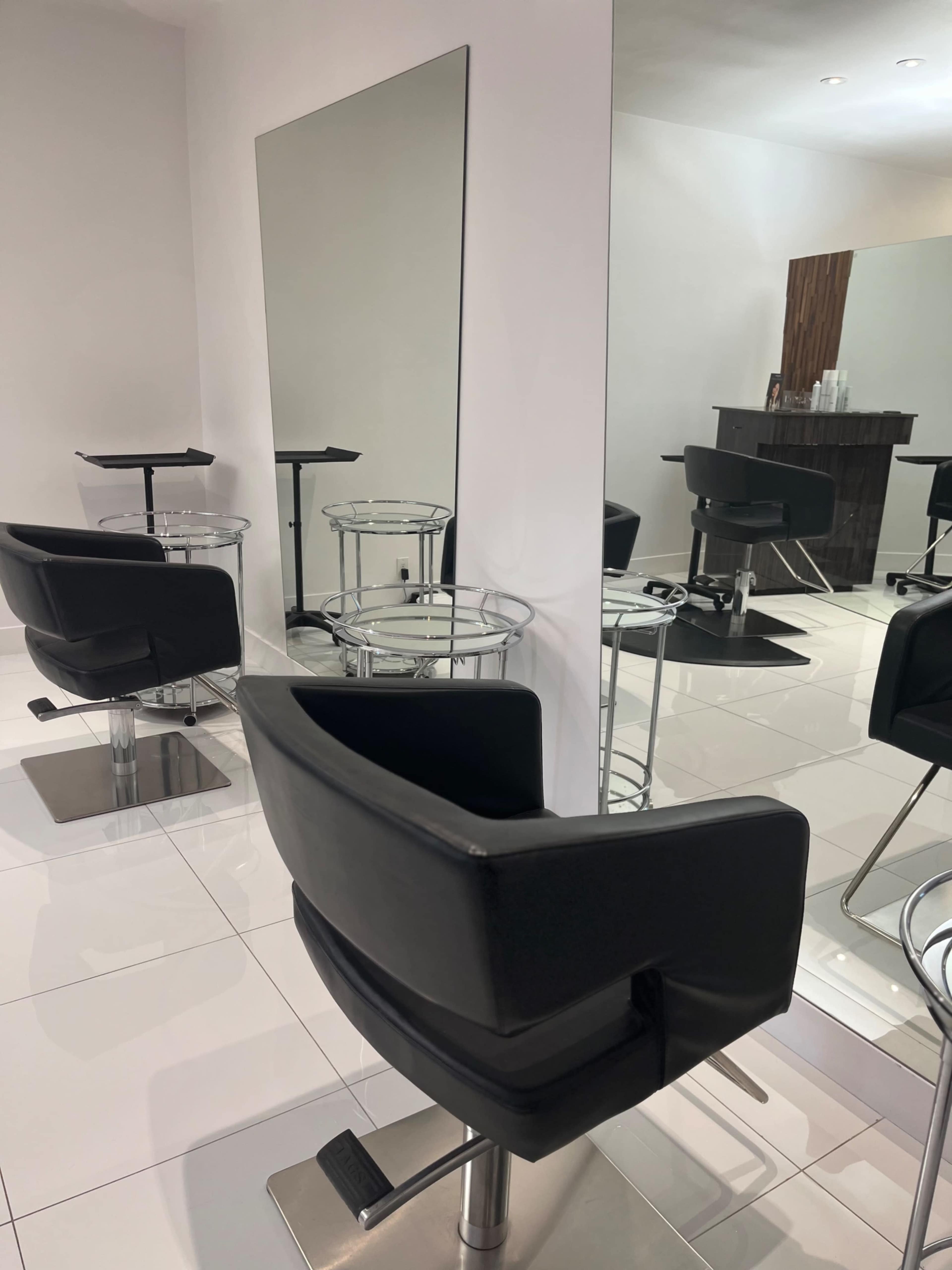 A modern salon interior with black reclining chairs and mirrors arranged against white walls and tiled floors.