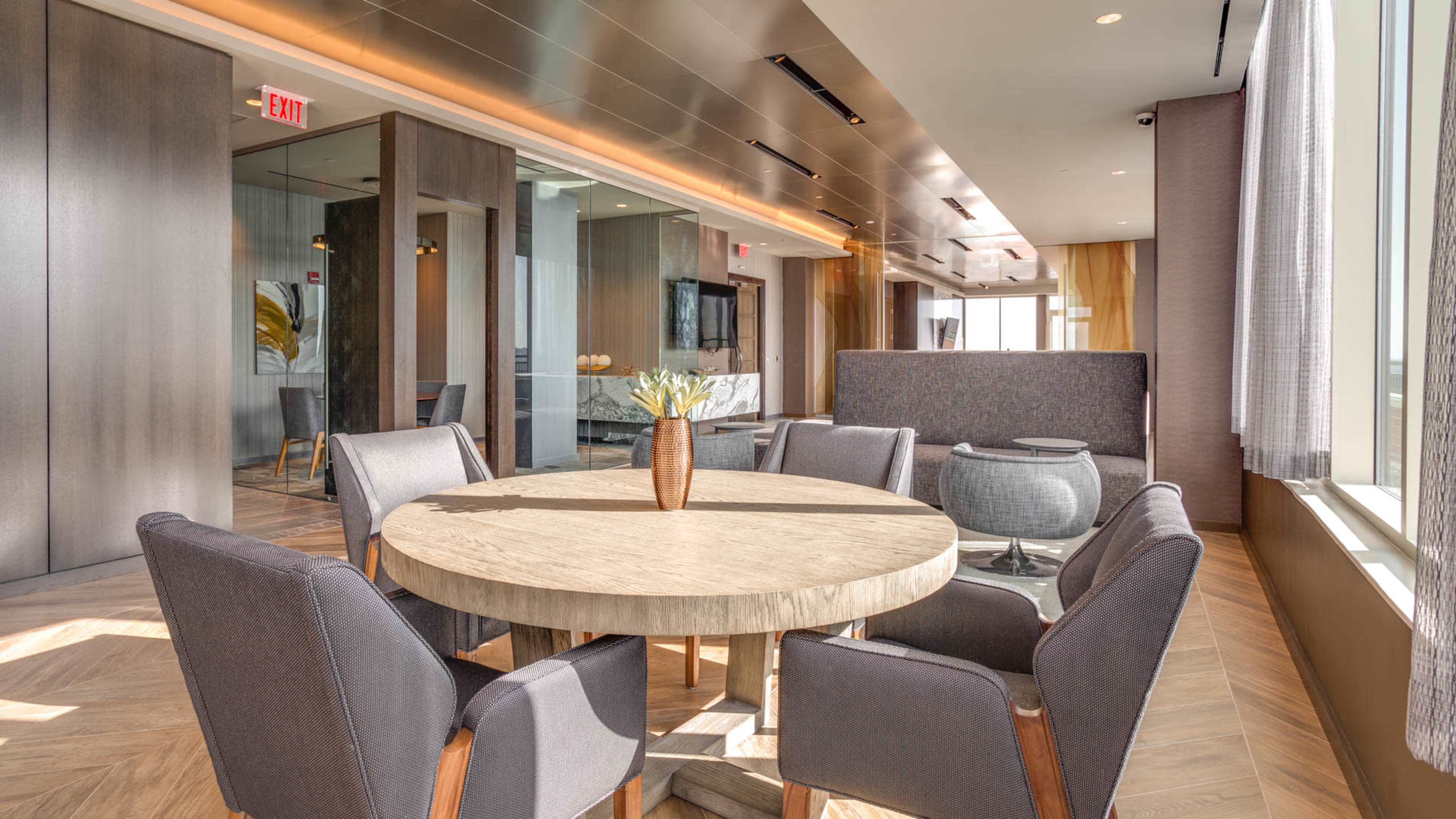 A modern lounge area features a round wooden table surrounded by gray upholstered chairs, with large windows providing natural light.