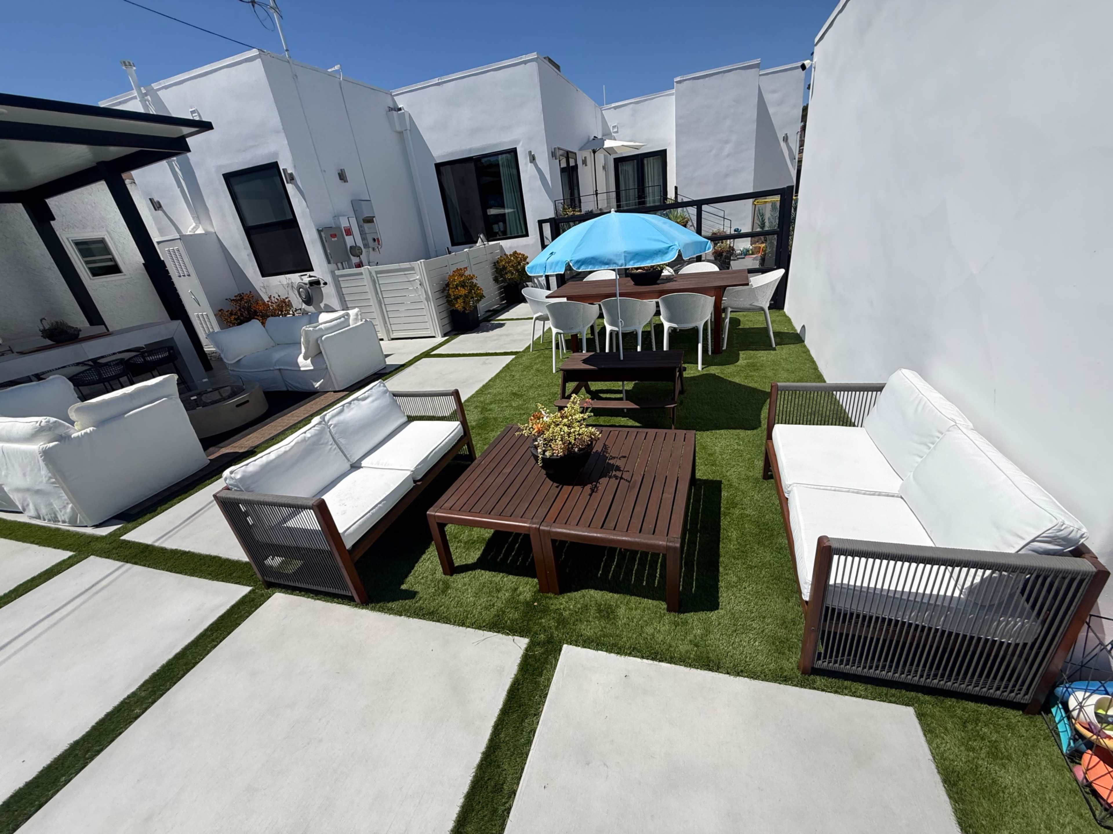 The image shows a modern outdoor patio with two seating areas, a central table, an umbrella, and a dining set on grass-like turf and concrete pathways.