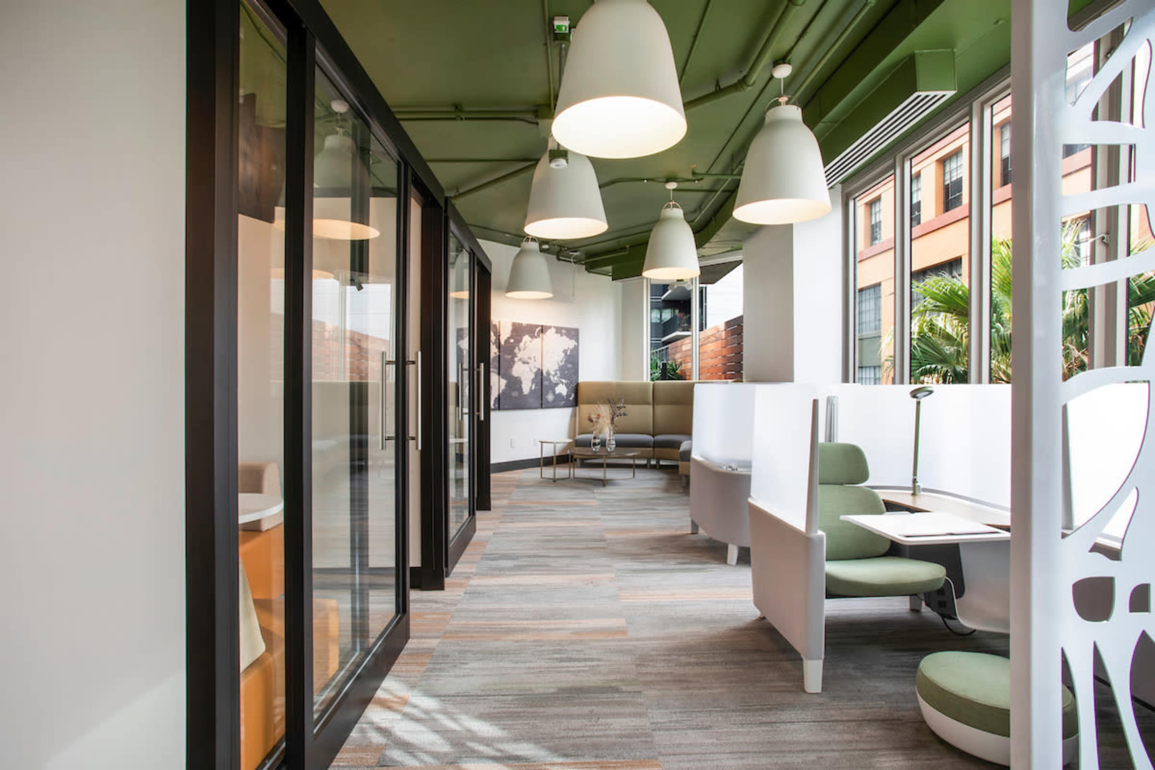 The image shows a modern office space featuring a series of workstations and seating areas illuminated by pendant lights and surrounded by large windows.