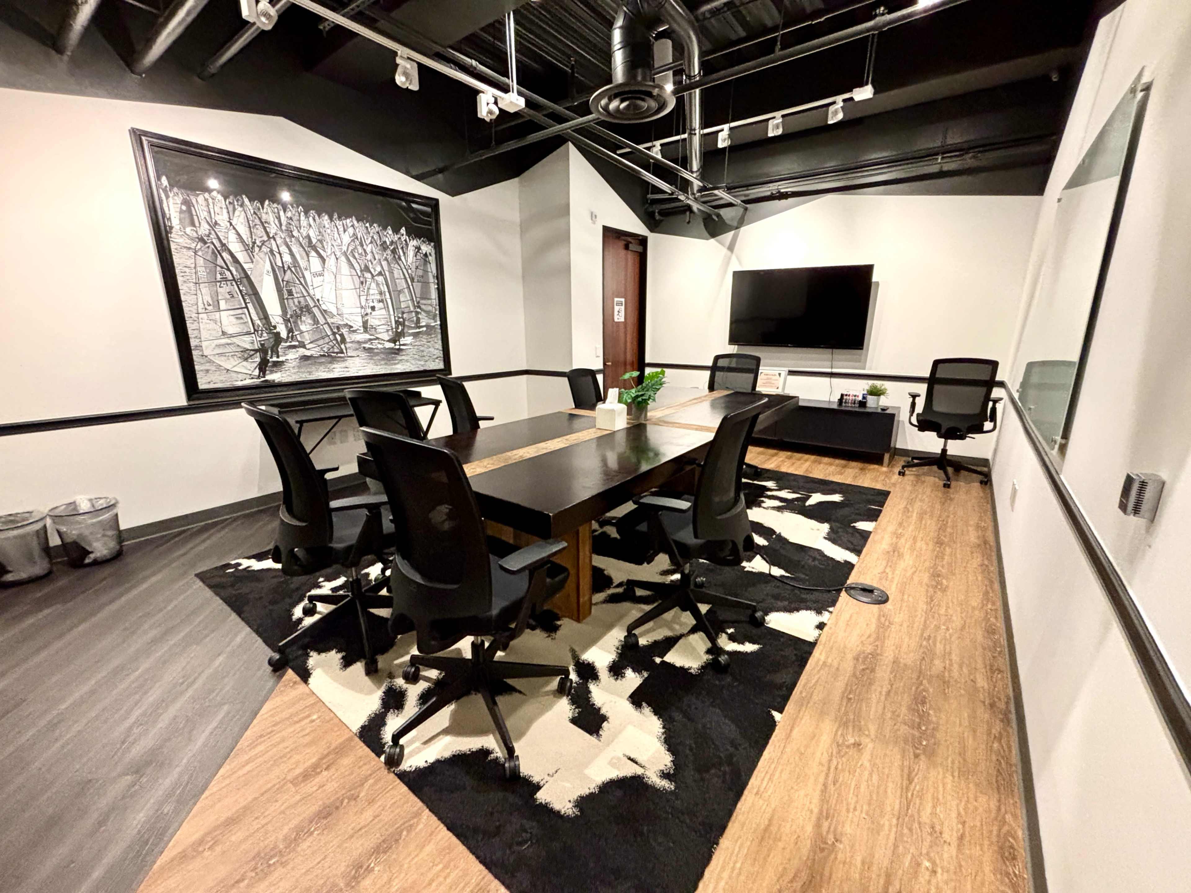 The image shows a modern conference room with a large table surrounded by black chairs, a wall-mounted television, and a black-and-white artwork.