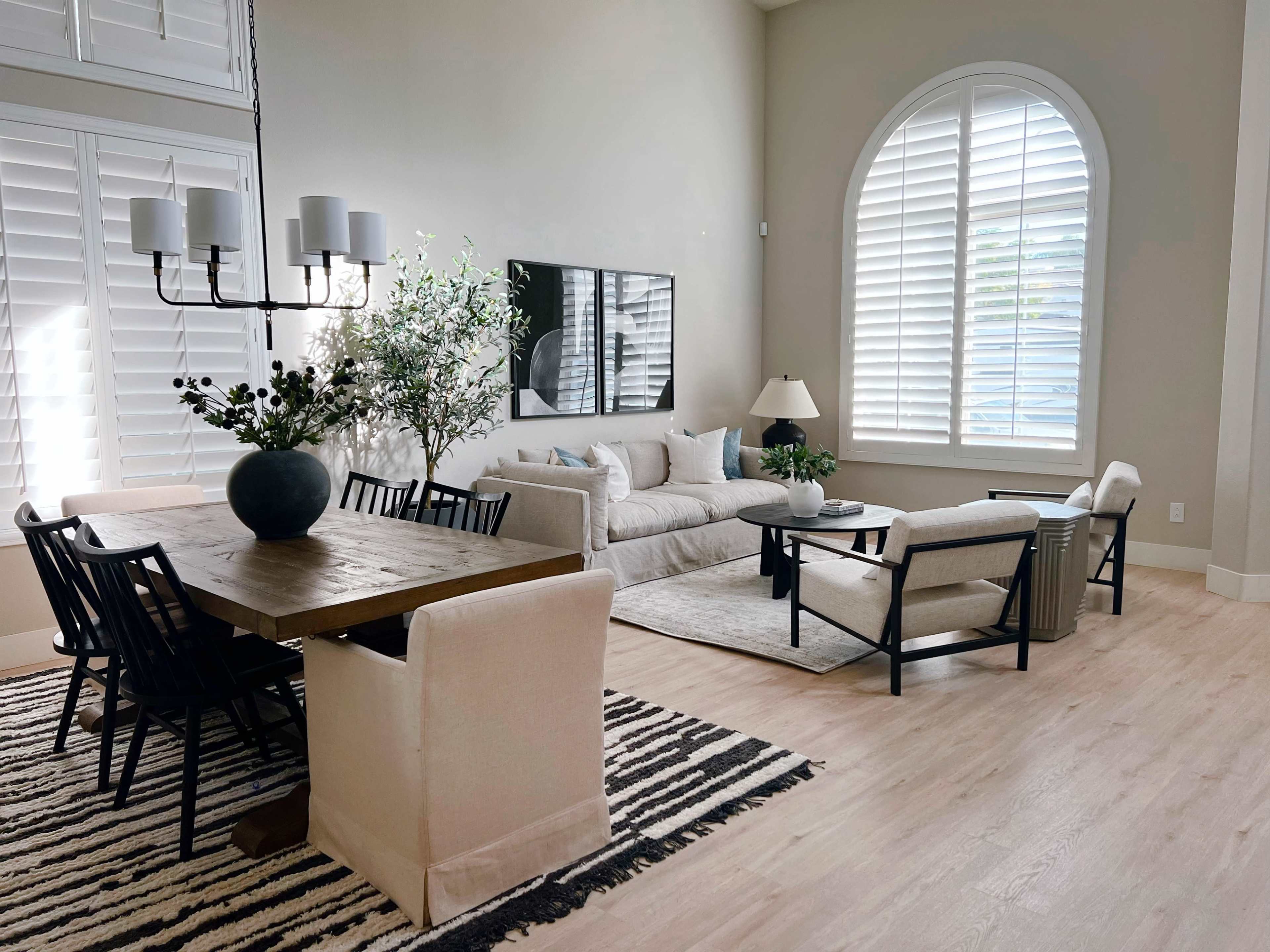 A modern living room with a light-colored sofa, a dining table, and large windows with plantation shutters.