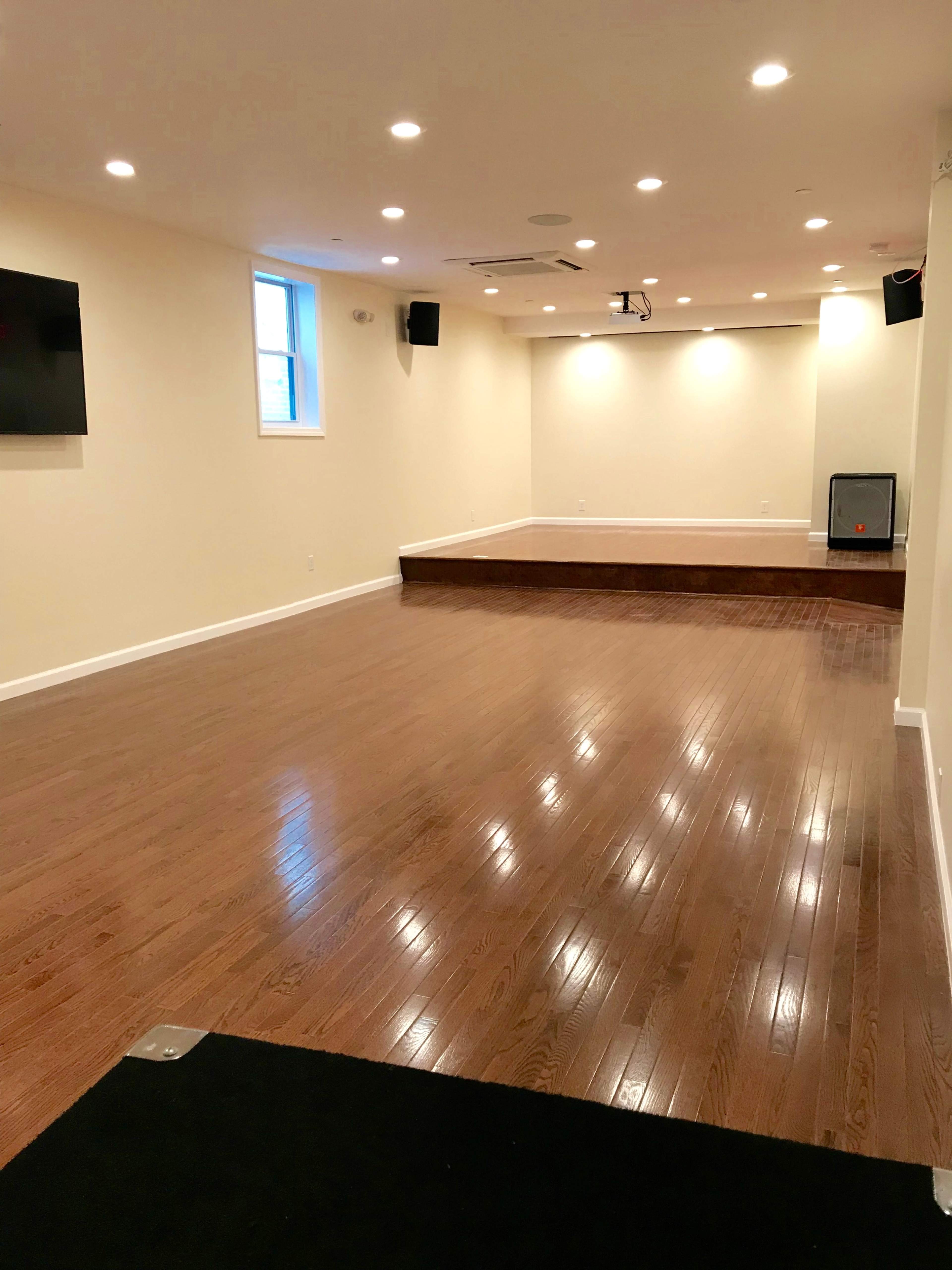 A spacious, empty room with polished wooden floors, bright overhead lights, and a small stage at one end.