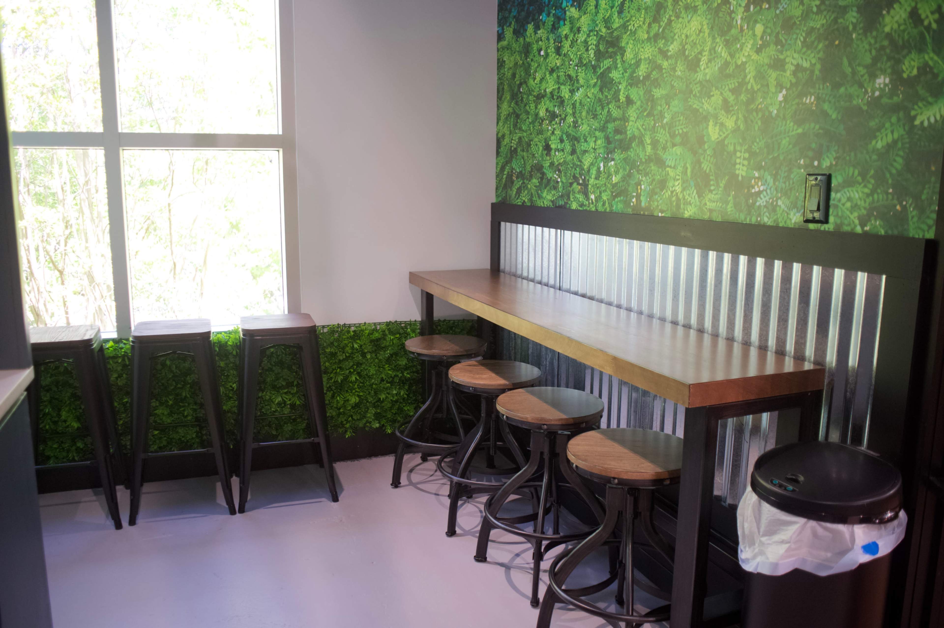 A minimalist dining area features a wooden counter with metal stools and a green plant-themed wall beside a large window.