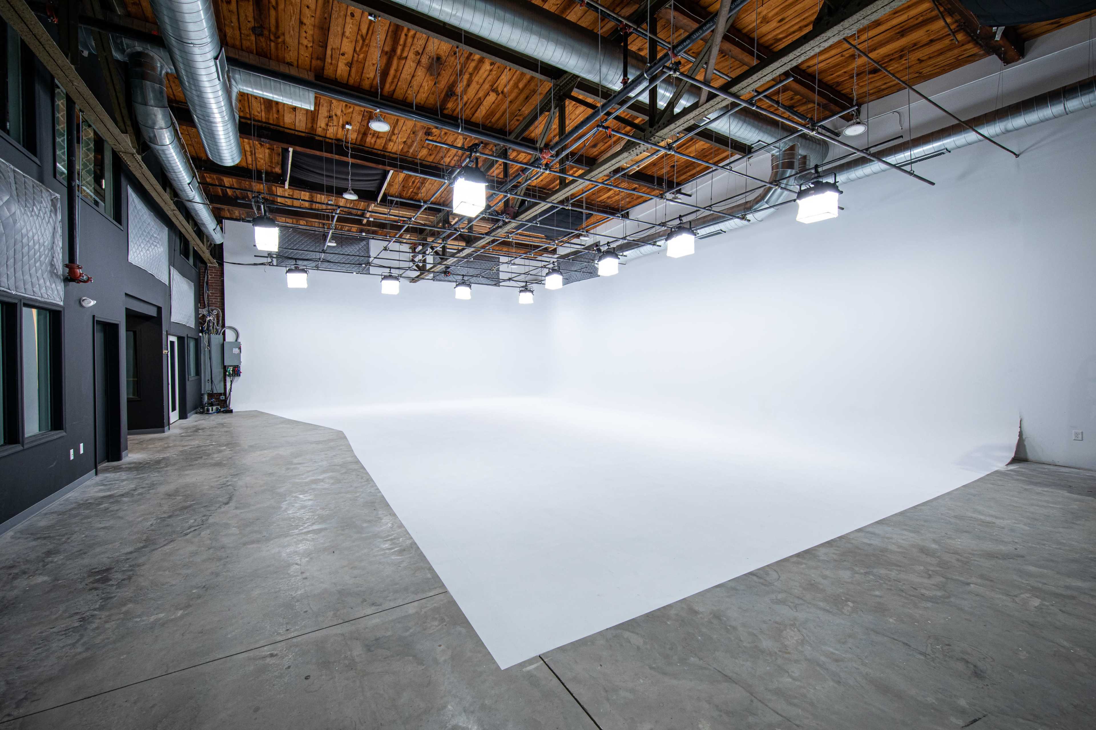 A spacious, empty studio with a white backdrop, wooden ceiling beams, and overhead lighting.
