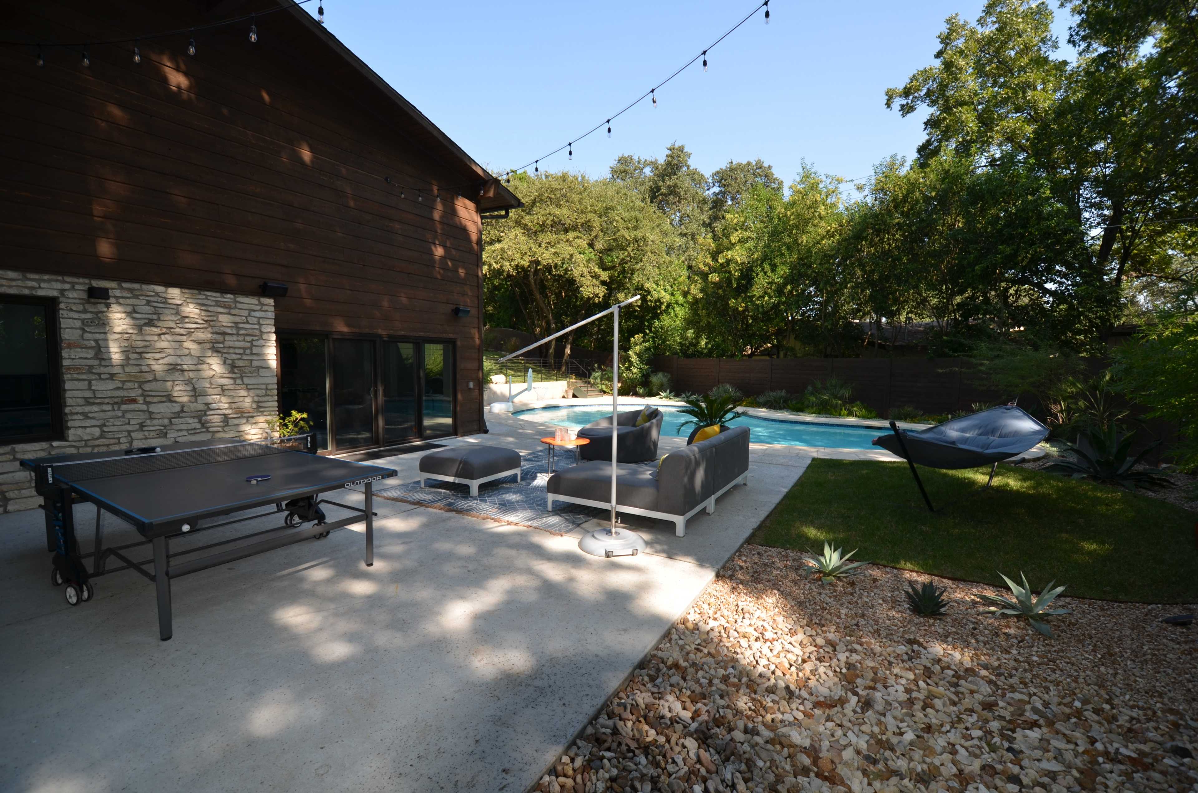 The image shows a backyard patio with a ping pong table, outdoor seating area, and a swimming pool surrounded by greenery.