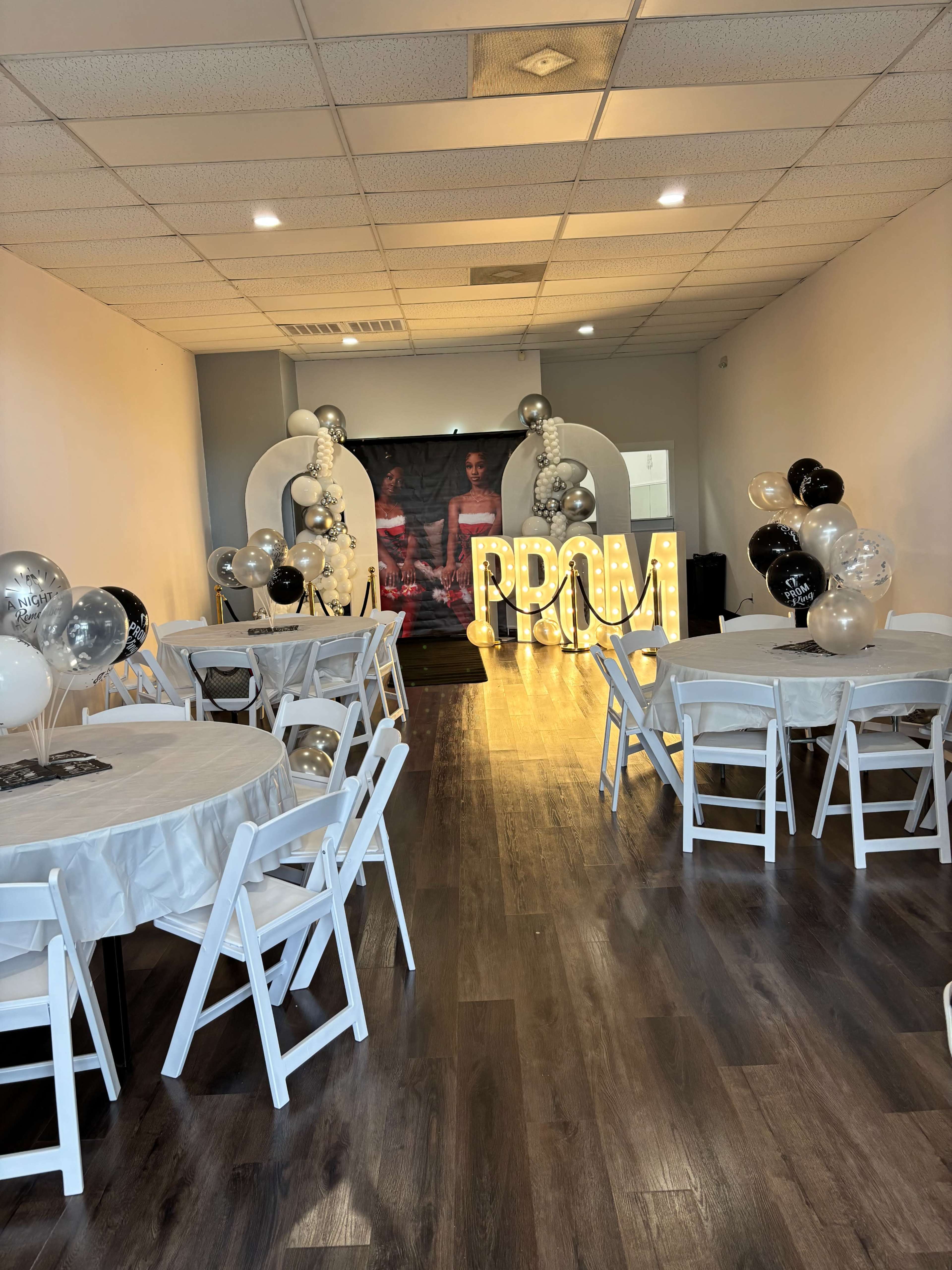 The image shows a decorated event space set up for a prom, featuring tables with white tablecloths, black and silver balloons, and illuminated letters spelling "PROM" at one end.