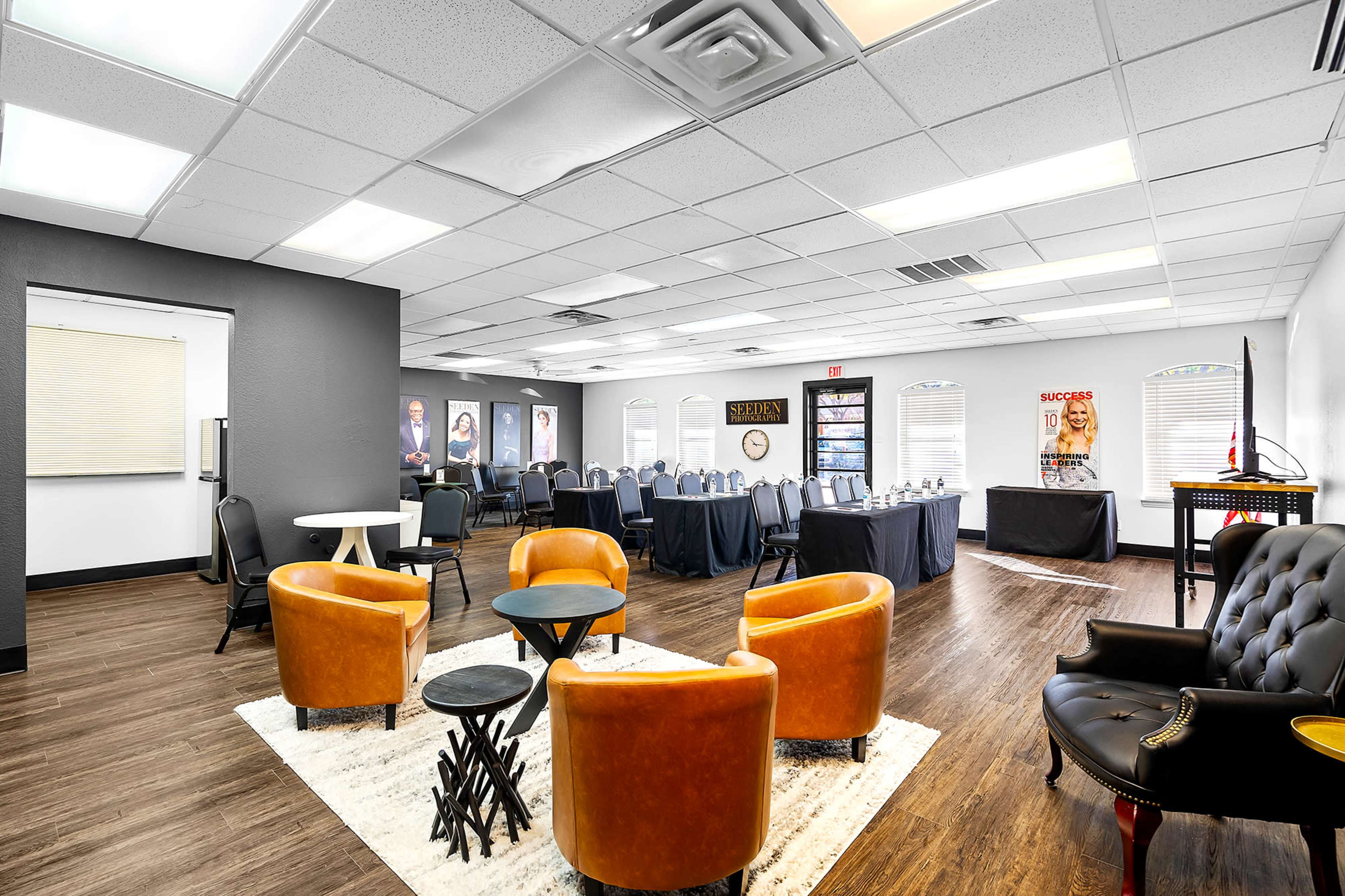 A modern event space with a seating area featuring orange chairs and a table, alongside a setup for tables arranged for a gathering in the background.