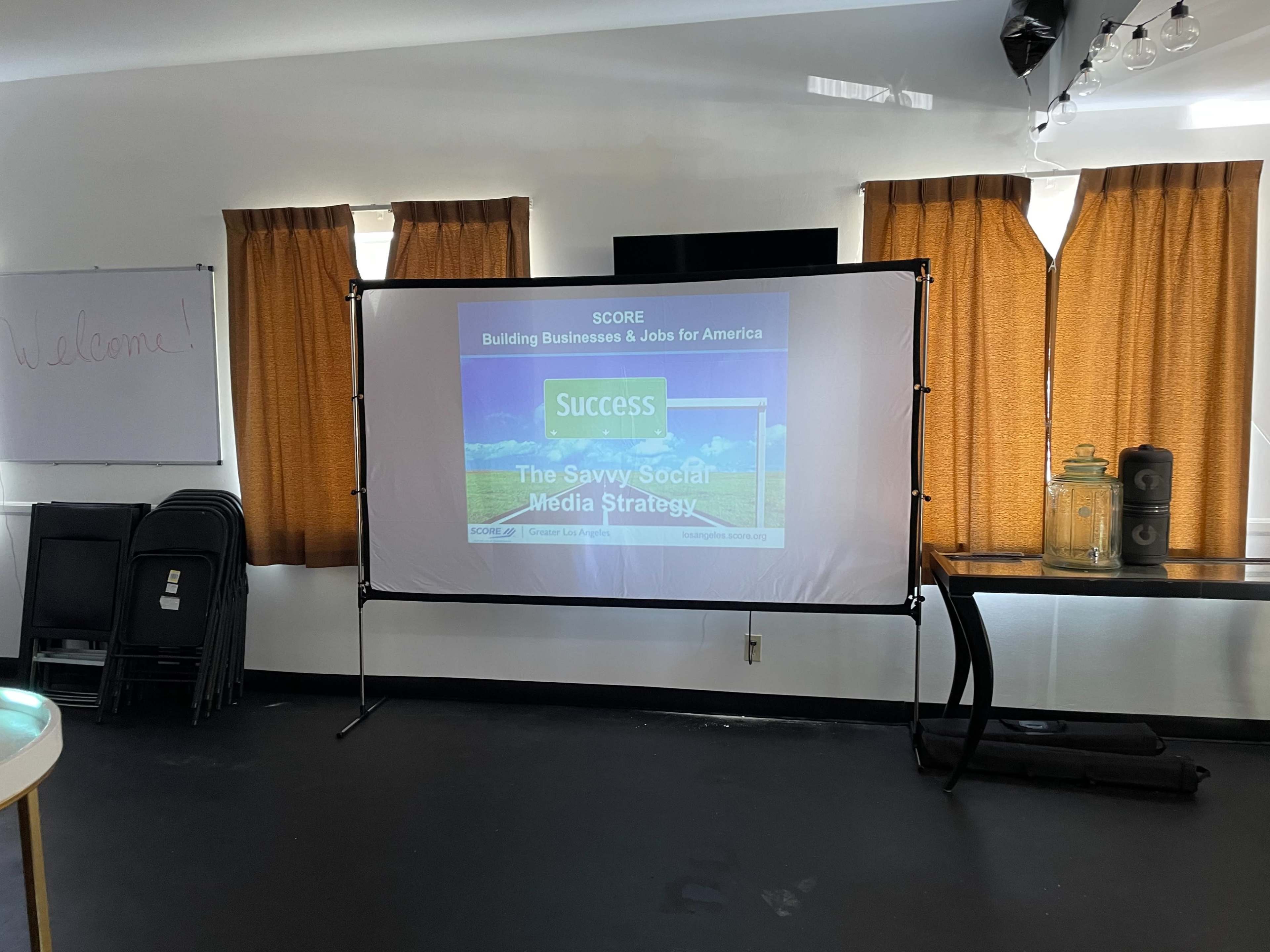 A projector screen displays a presentation titled "The Savvy Social Media Strategy" in a room with orange curtains and folded chairs stacked in the corner.