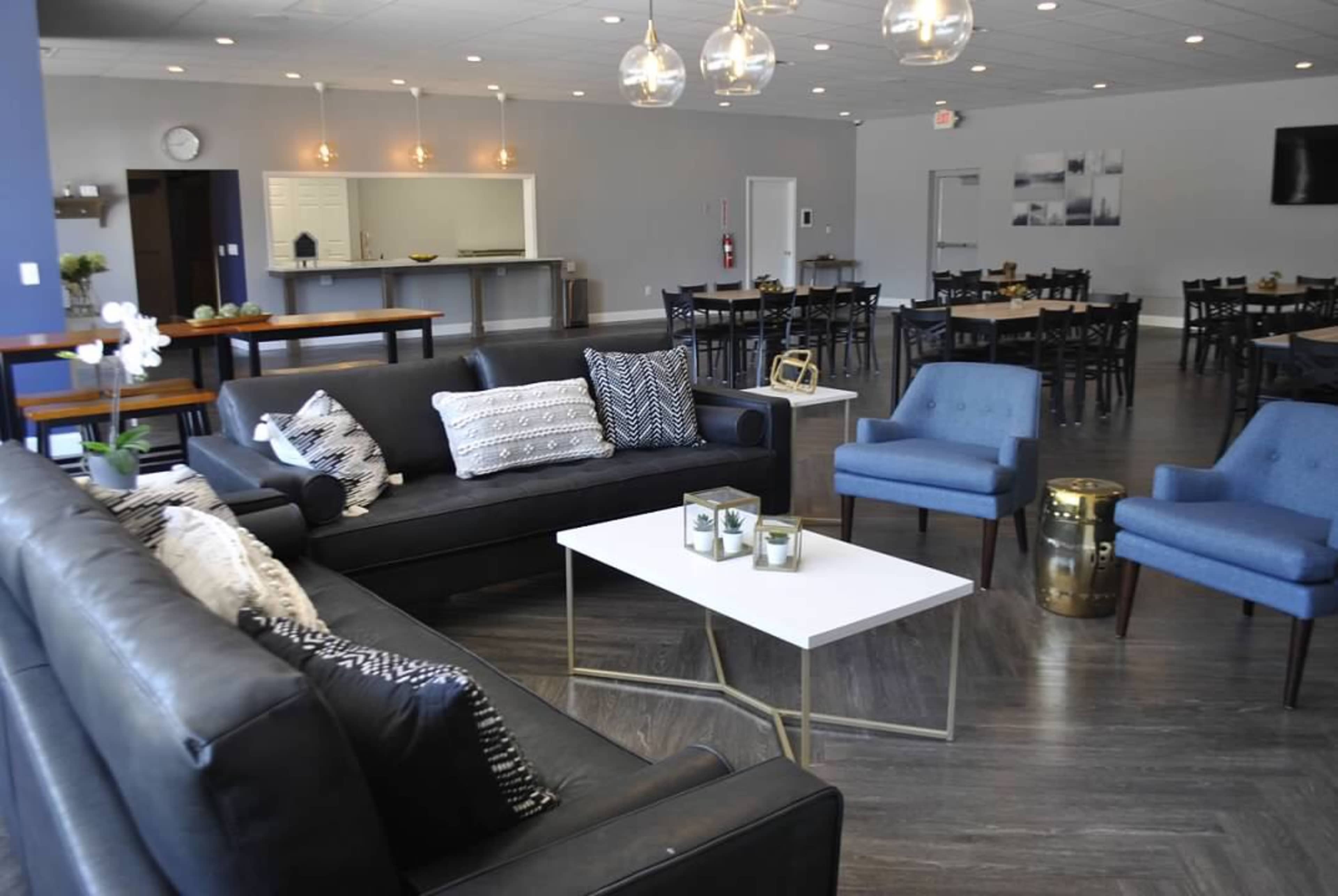 A modern indoor communal space featuring black sofas, blue chairs, and a white coffee table, with a dining area in the background.