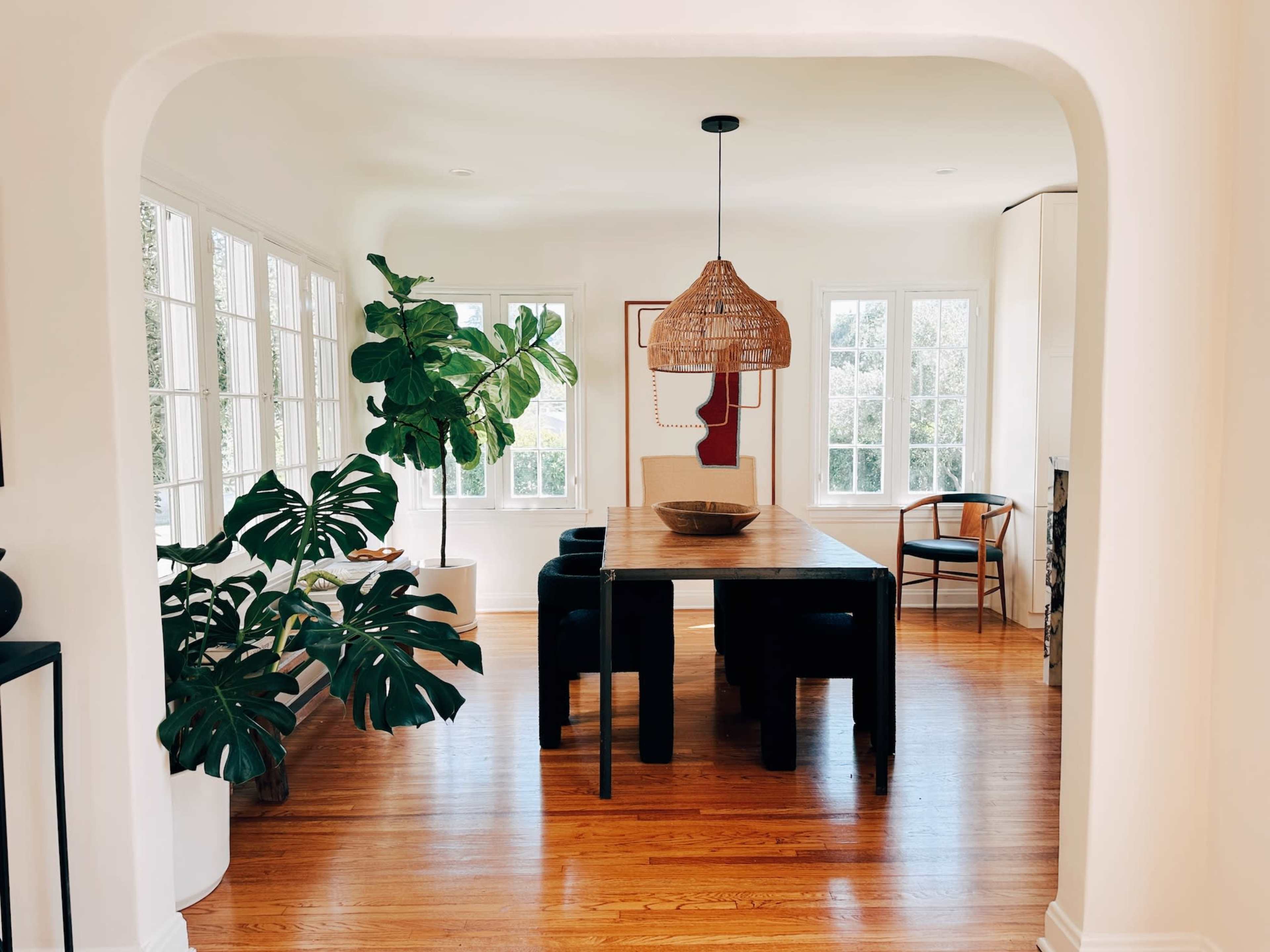 A dining area features a wooden table surrounded by black chairs, with large windows and a potted plant nearby.