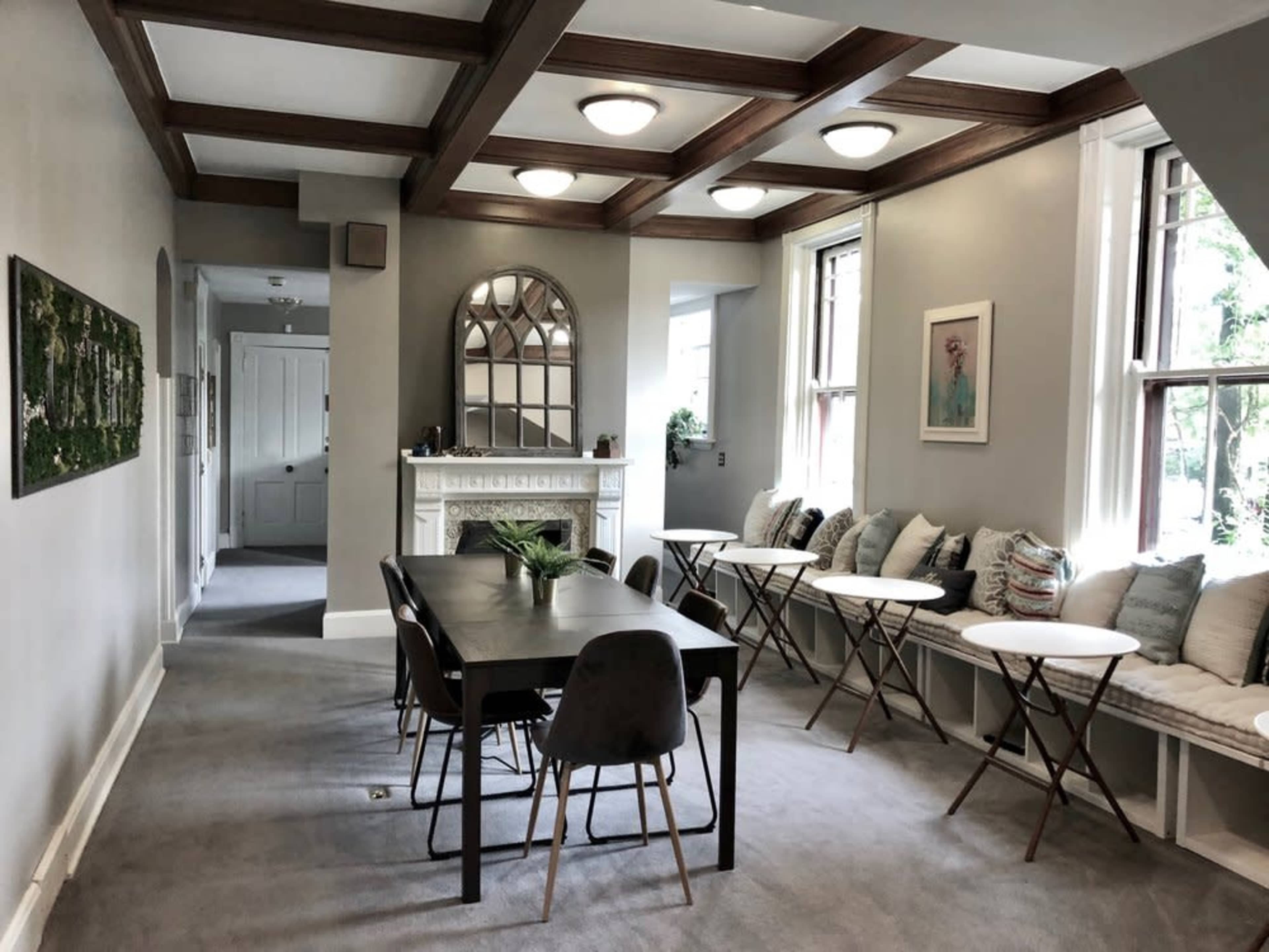 The image shows a spacious room with a long table, several small round tables, a fireplace, and large windows, featuring a neutral color scheme and a modern design.