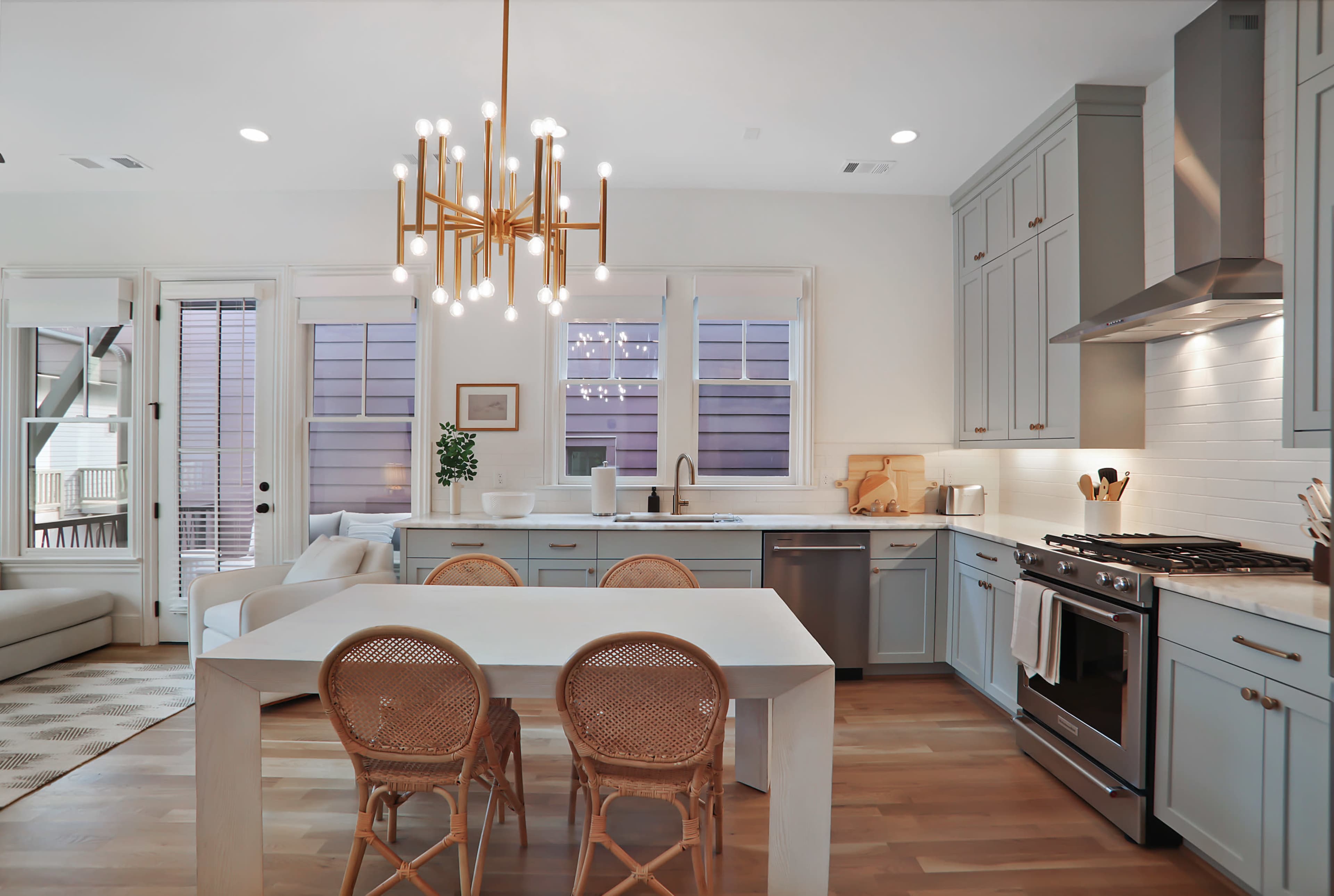 A modern kitchen features blue cabinets, a white dining table with rattan chairs, and a gold chandelier.