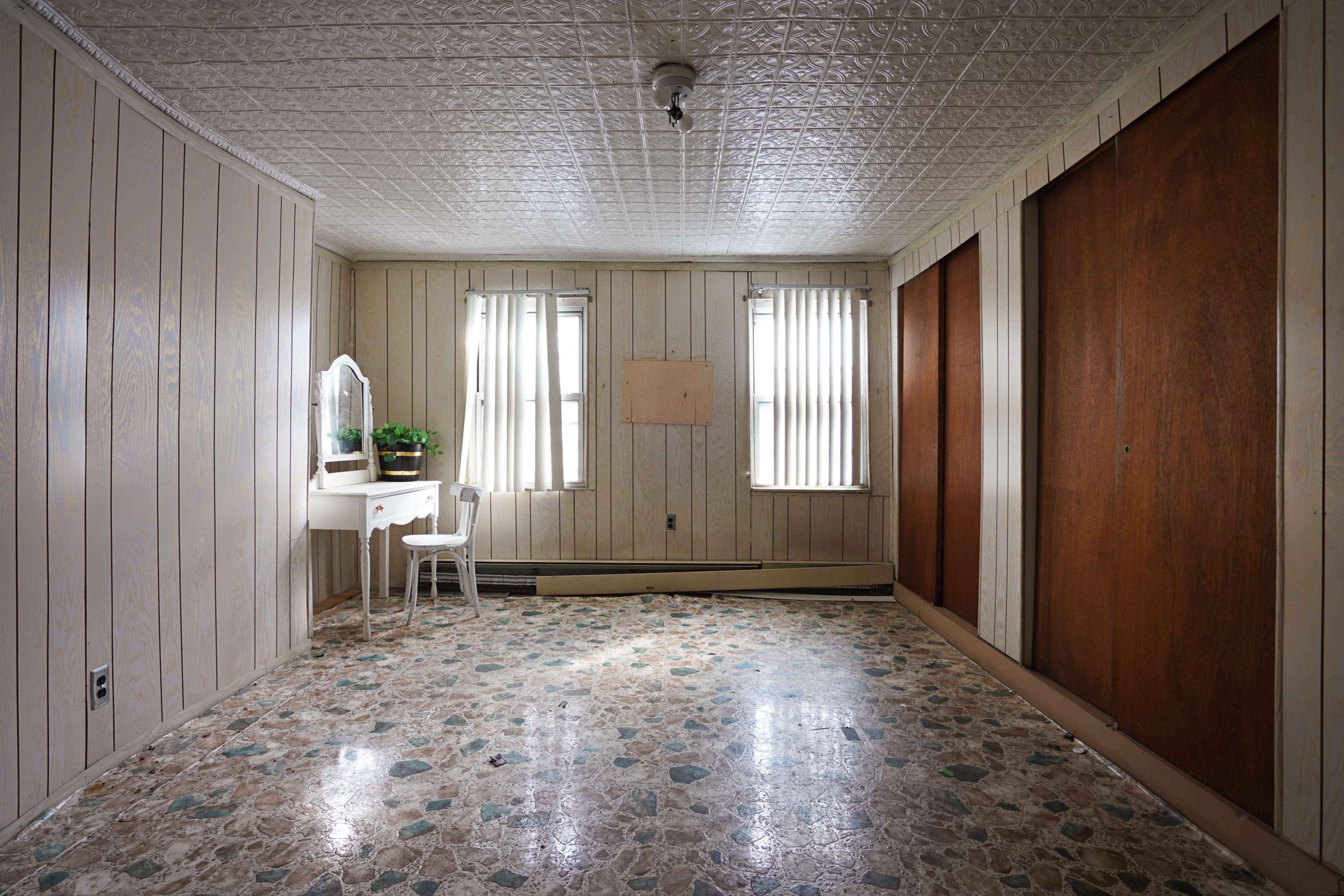 A sparsely furnished, empty room with wooden paneling, two windows with vertical blinds, and a small white vanity against one wall.