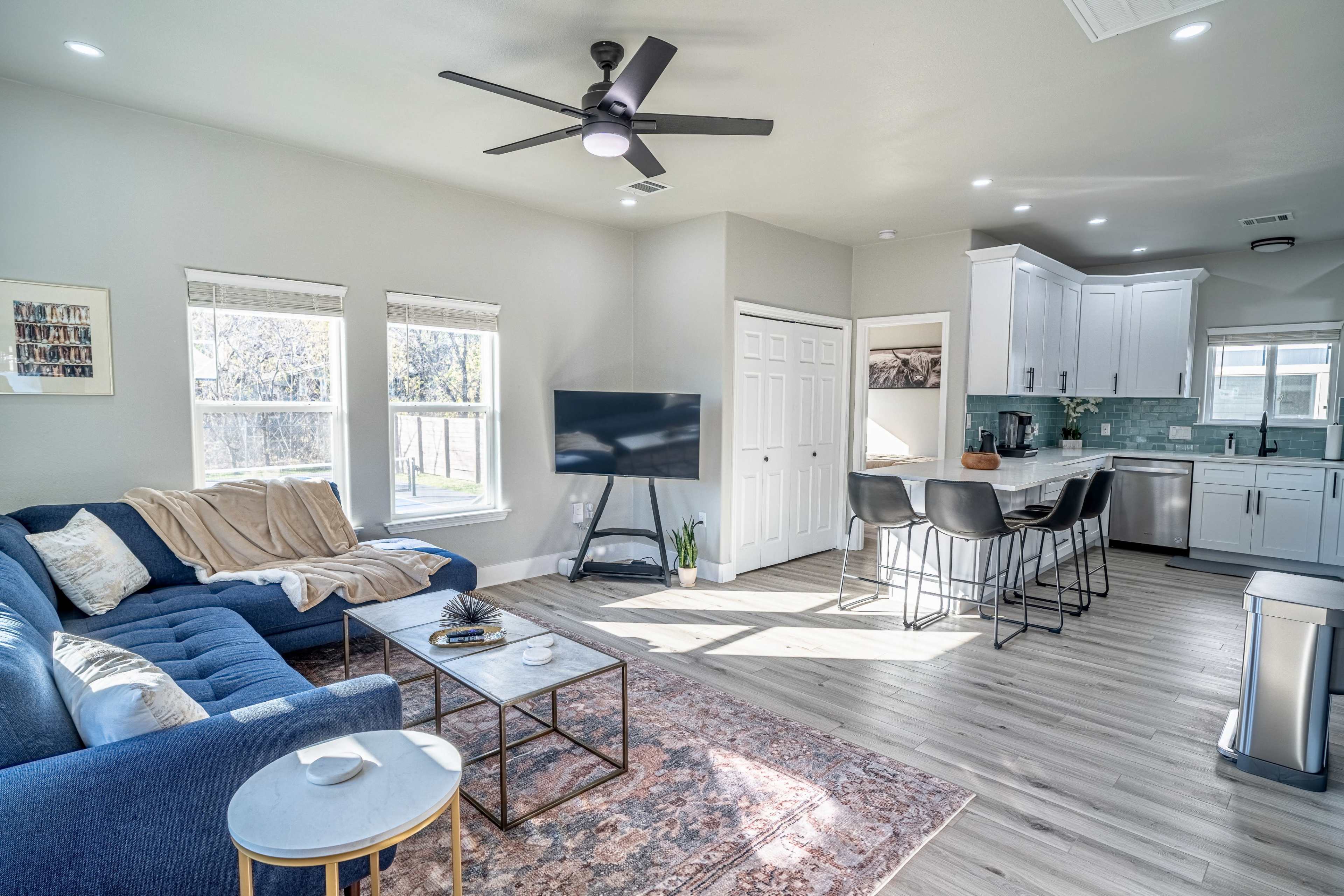 The image shows a modern living room with a blue sofa, a coffee table, and a flat-screen TV, adjacent to a kitchen area with white cabinets and bar stools.