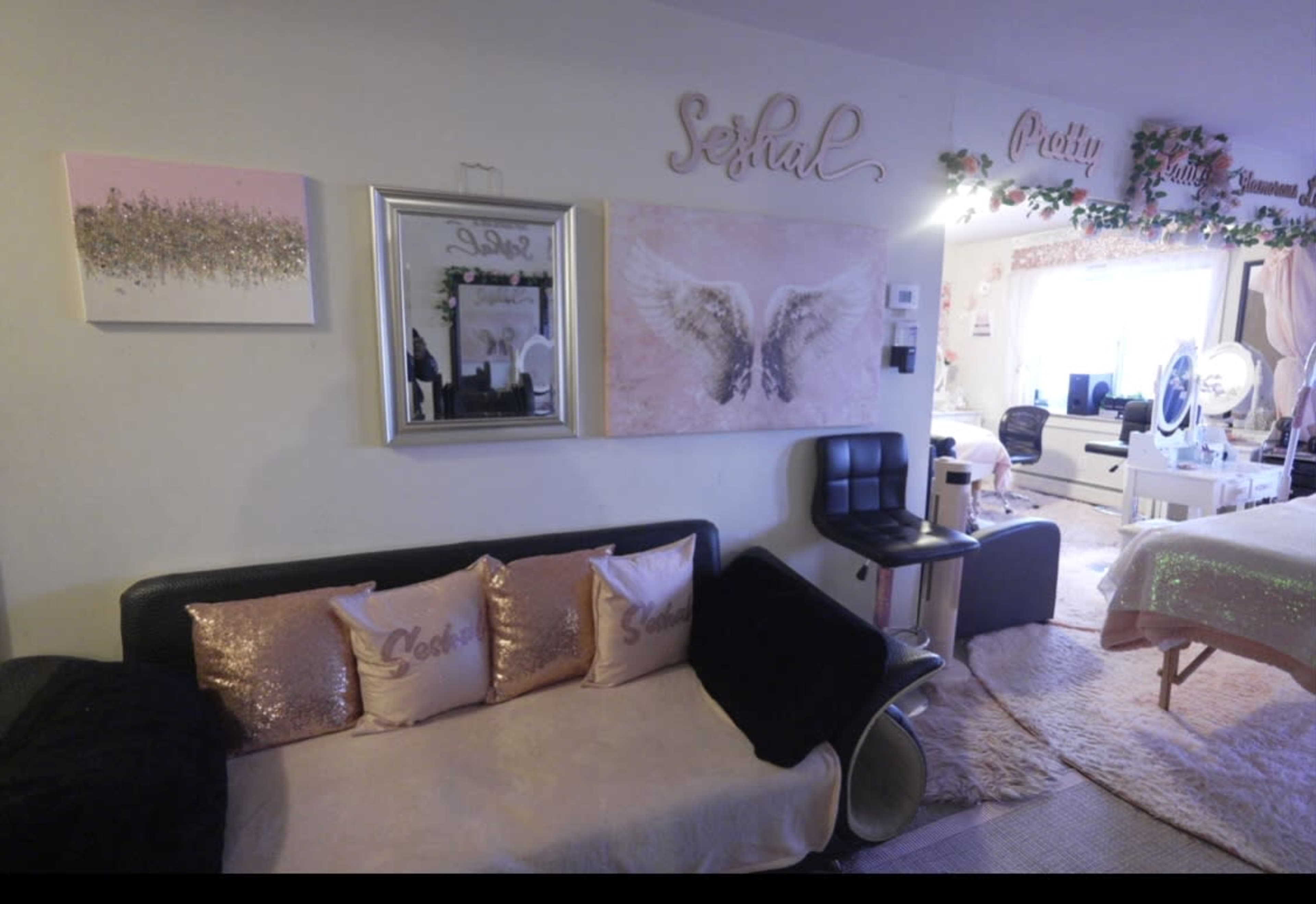 A cozy room with a black sofa, decorative pillows, pink artwork on the walls, and a well-lit area featuring a makeup station and a bed.