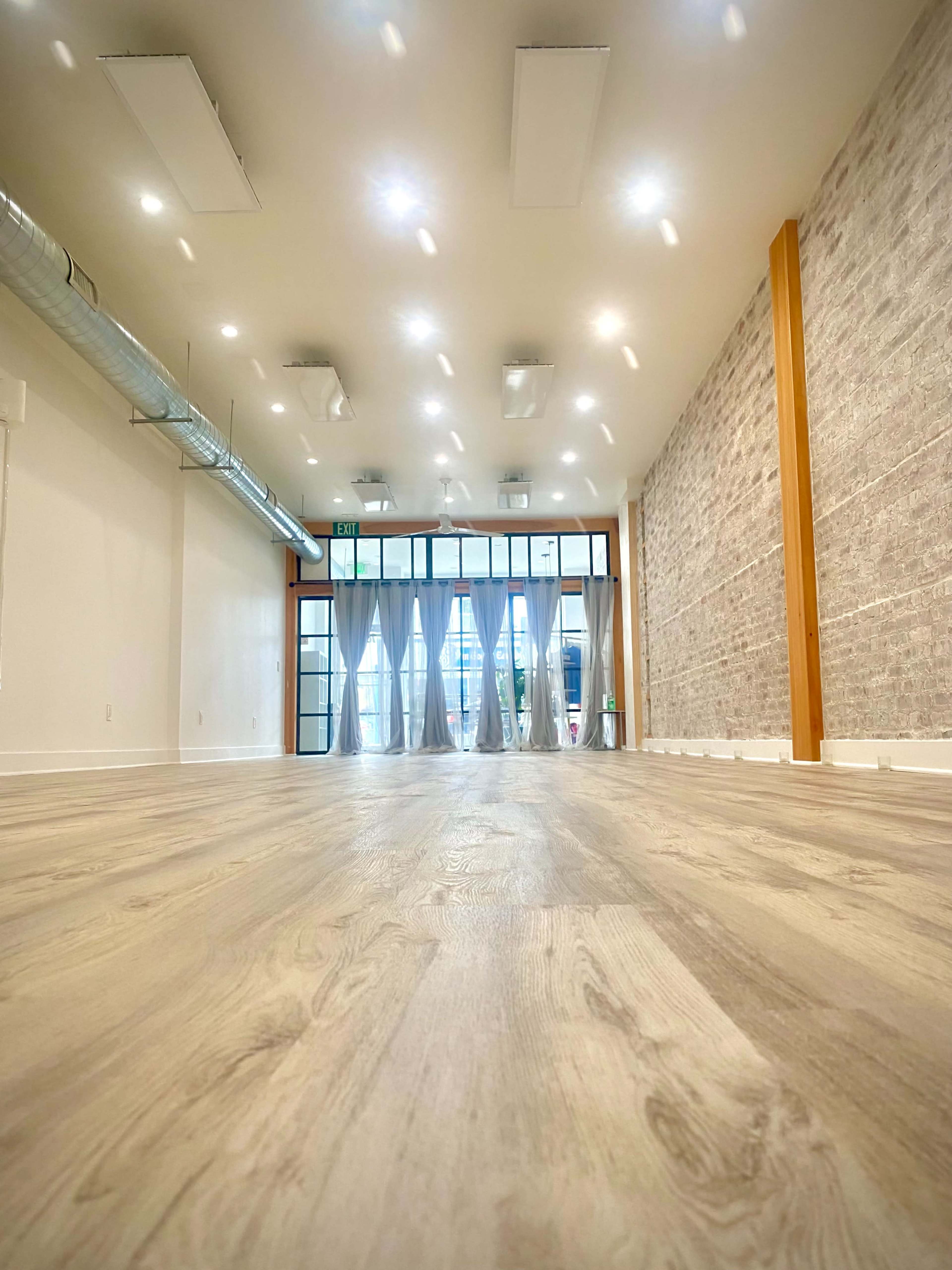 A spacious interior with light-colored wooden flooring, modern lighting fixtures, and large windows along one side, providing a view of the exterior.