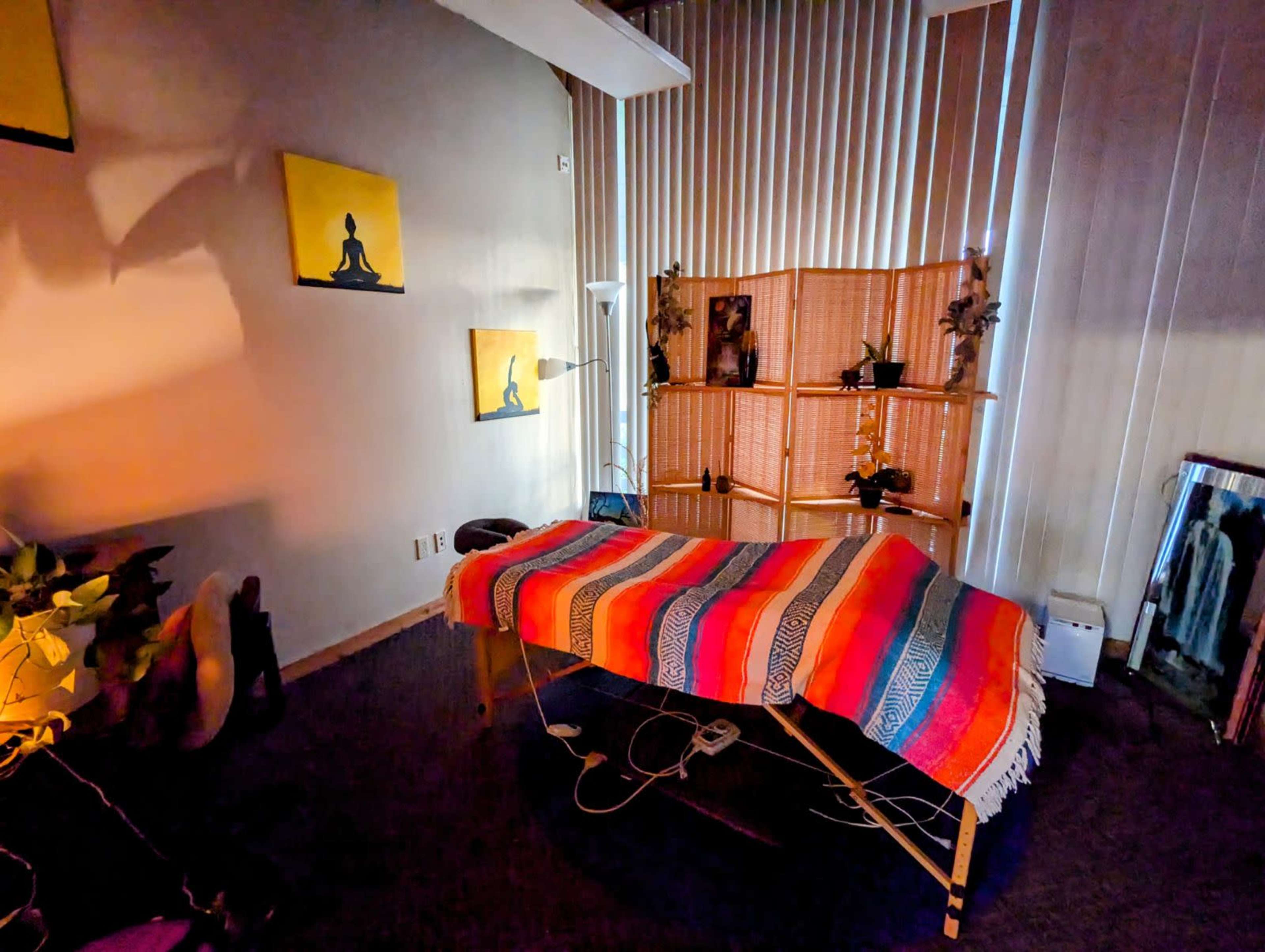 The image shows a massage table covered with a colorful blanket in a softly lit room decorated with framed artwork and plants.